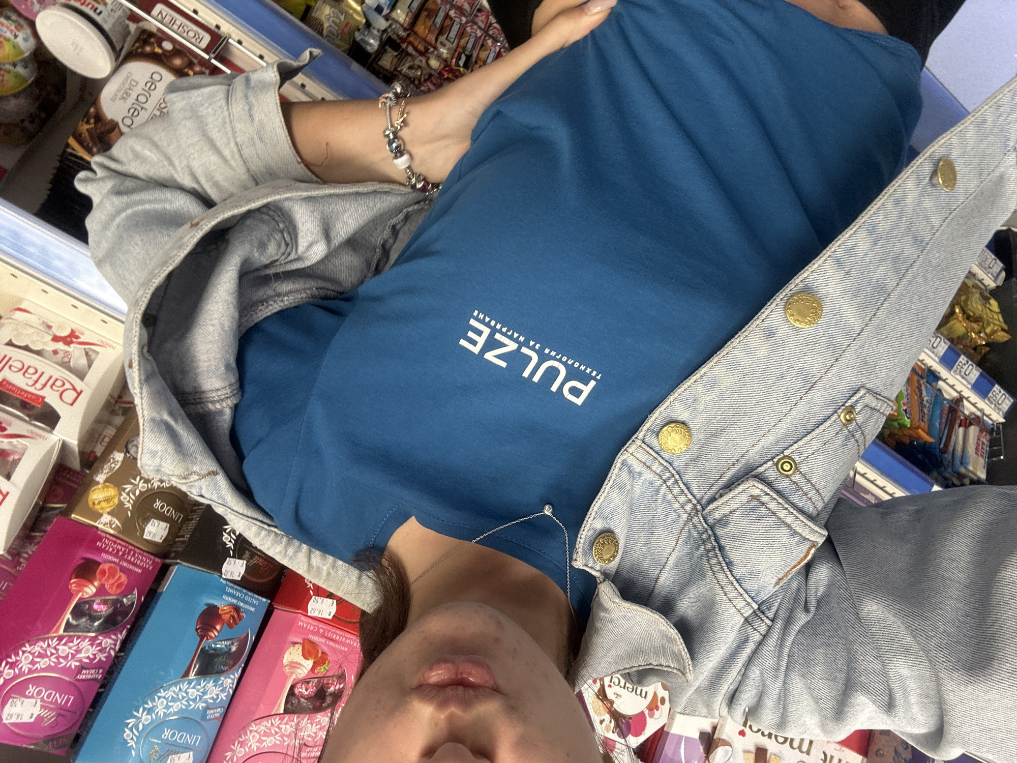 A young woman, seen from the chest up, stands in an aisle of a store. She is wearing a blue t-shirt with a white logo that reads "PULZE" and smaller text below it. Over the t-shirt, she has on a light-wash denim jacket, unbuttoned. The jacket has two gold buttons visible on the right side. She is wearing a delicate silver necklace with a small diamond pendant. The background is a store shelf filled with various chocolate boxes, including brands like Lindt, Merci, and Raffaello. The lighting suggests an indoor retail environment.