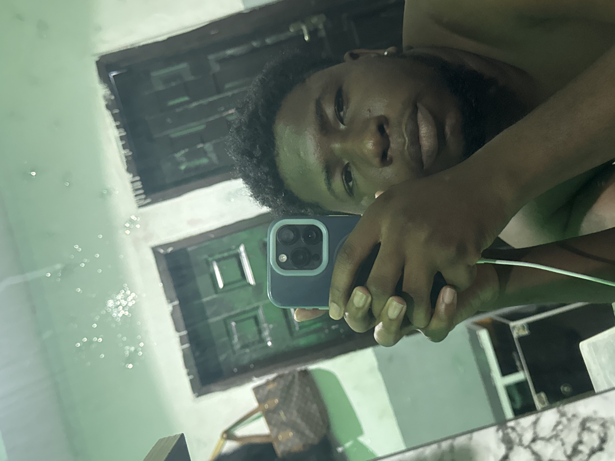 A man takes a selfie in a mirror. The man has dark skin, short curly black hair, and a beard. He is holding a blue smartphone in his right hand. A dark-colored Louis Vuitton bag sits in the background, along with a chair. The mirror reflects a window. The location is potentially Ipetumodu, Nigeria.