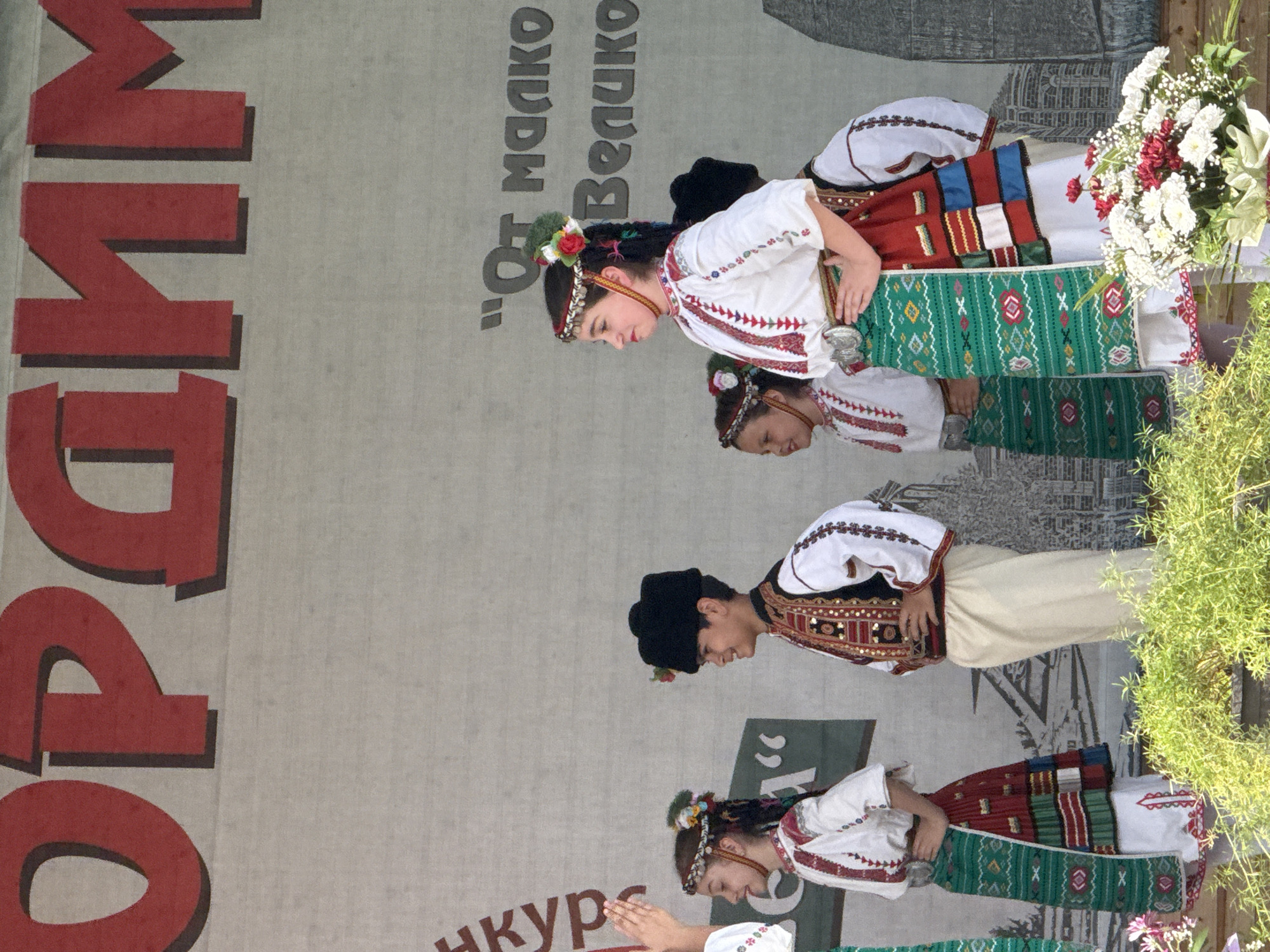 The image depicts a group of children in traditional Bulgarian folk costumes performing on a stage. In the foreground, several children, including girls in embroidered white blouses, colorful skirts, and headbands adorned with flowers, and a boy in a black fur hat, embroidered vest, and white pants, are the main subjects. They appear to be engaged in a dance or presentation, with some individuals looking to the side and others facing forward. The backdrop features a large banner with red lettering. Part of the banner reads "Орхим" in large red capital letters, and to the right, smaller text in black is visible, including "От Малко" and "Велико". The setting suggests an outdoor event, possibly a festival or cultural celebration, held in the city of Pordim, Bulgaria, given the prominent display of the city's name on the banner. Lush green foliage is visible at the bottom of the image, hinting at a natural or park-like environment.