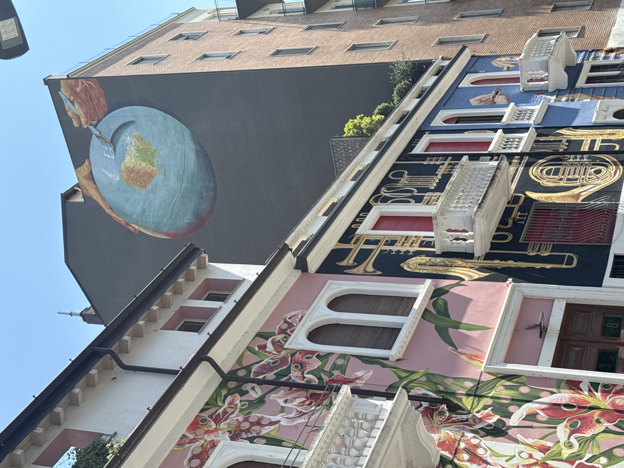This is an upward-angle, street-level photograph of building facades in Milan, Italy. The walls are covered in elaborate trompe-l'oeil paintings. The lower facade is pink, featuring flower paintings and architectural elements. Above, a navy blue facade presents golden musical instruments and more architectural details. A taller brick-colored building with a mural of two hands painting the earth on a plate is above. The sky is clear and blue.