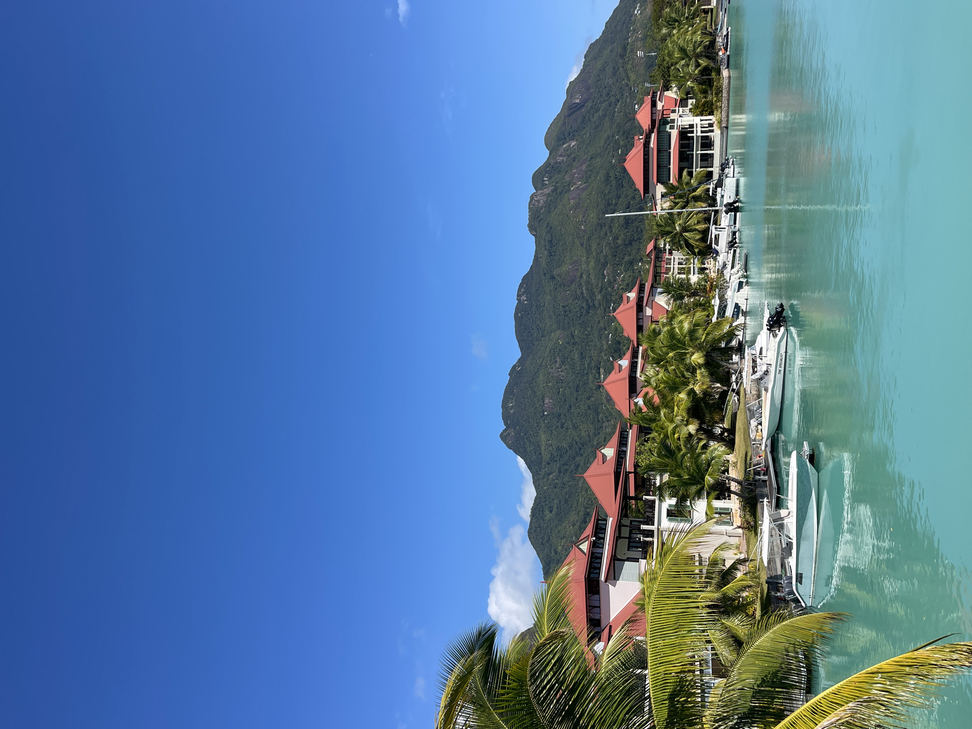 The image depicts a serene waterfront scene in Victoria, Seychelles. In the foreground, the turquoise water of a harbor is calm, with several white motorboats docked along a wooden pier. Lush green palm trees frame the left side of the image. Behind the boats, a row of white buildings with distinctive red-tiled roofs, characteristic of resorts or residential complexes, stretches along the waterfront. These structures are nestled amongst more palm trees. In the background, a lush, green, mountainous landscape rises under a clear blue sky dotted with a few white clouds. The overall impression is one of tropical tranquility and upscale coastal living.