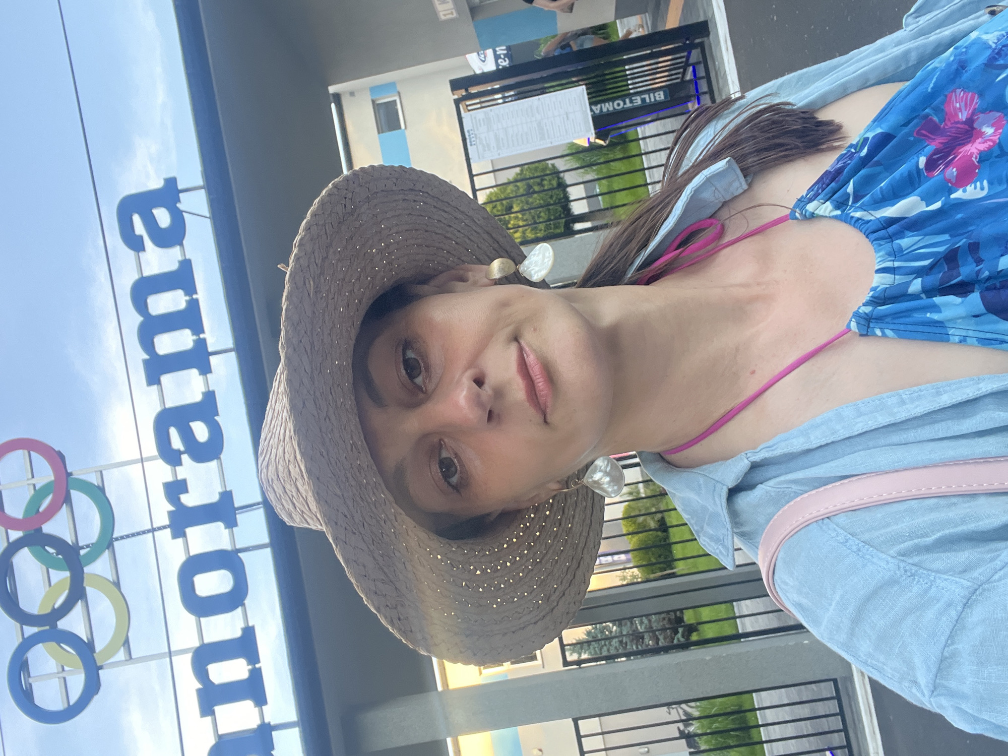 A woman wearing a straw hat and a blue linen shirt over a blue floral swimsuit takes a selfie in front of a building with the word "Panorama" in large blue letters above the entrance. Above the word are the Olympic rings, indicating a possible connection to the Olympics. She is also wearing large, white, shell-like earrings and a pink necklace. A light pink purse strap is visible over her shoulder. A metal fence is in front of her, with a sign to her right that appears to be a ticket booth or information kiosk. Trees and a building are visible in the background. The setting appears to be outdoors, possibly a public venue or attraction.