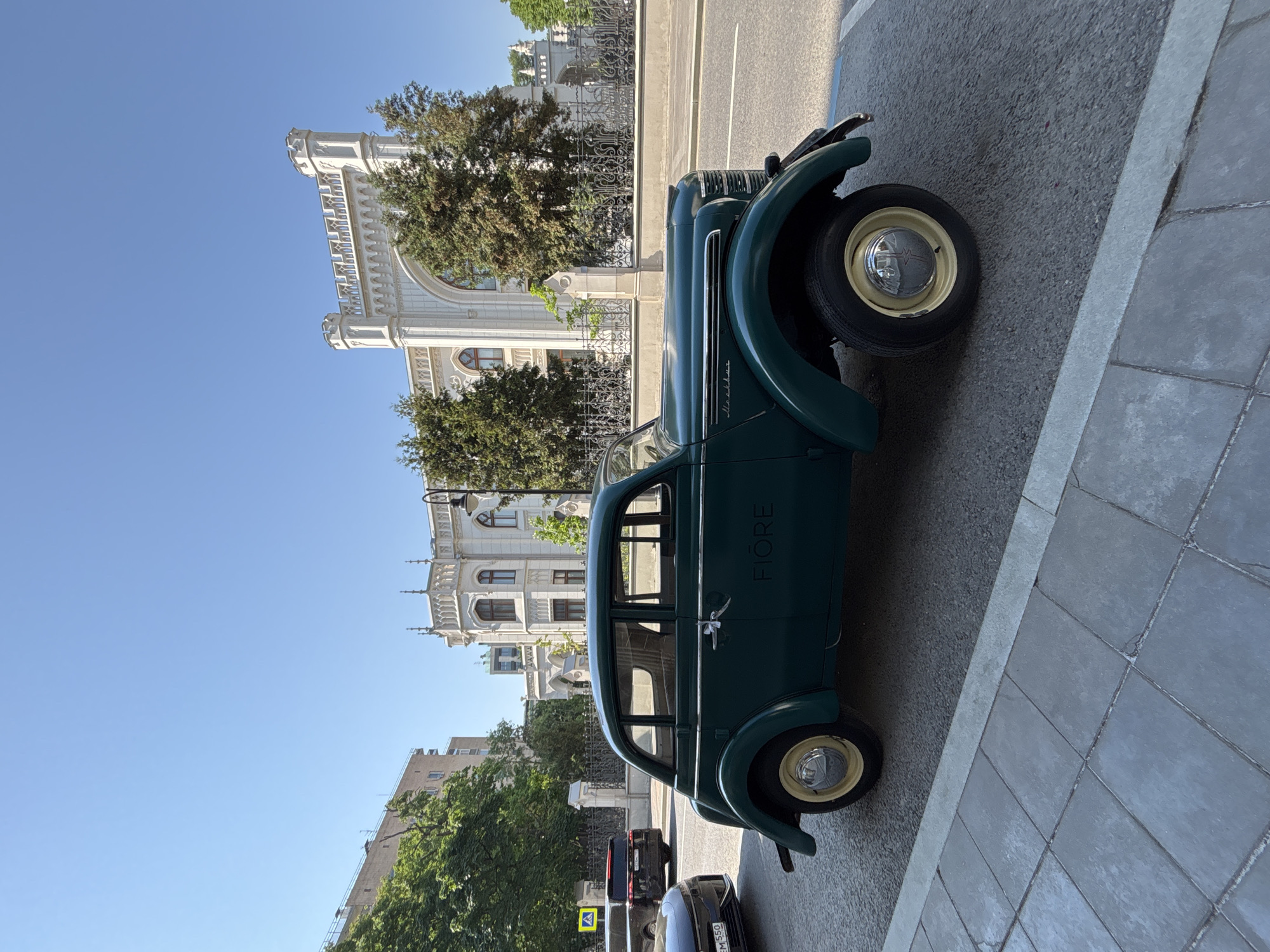 The image shows a vintage green car parked on the side of a street in Moscow, Russia. The car has the word "FIORE" painted on its side. The architectural style of the building behind the car suggests that the location may be near the State Kremlin Palace. Other cars are parked behind the vintage car. The sidewalk is paved with gray square tiles. The sky is clear and blue. The time of day appears to be during daylight hours. There are trees visible in the background.