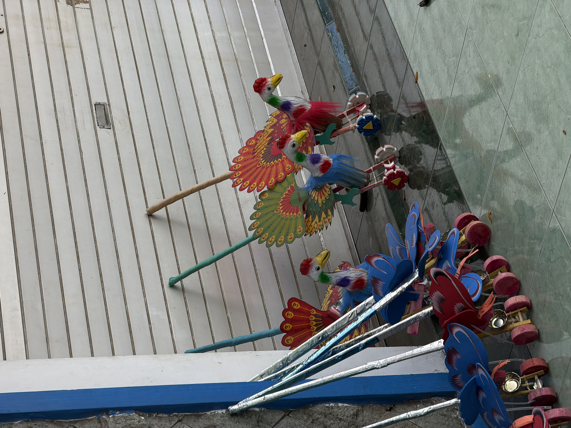 In Curahsawo, Indonesia, a row of colorful, handmade bird toys on wheels stands outside a building with a white, corrugated metal roll-up door. The toys, made of wood and paper, depict birds with brightly colored plumage, featuring red, blue, green, yellow, and orange accents. Each toy is mounted on a wooden stand with red and blue wheels, resembling a wheeled cart. Several silver poles, likely part of the toy construction or display, lean against a blue and white pillar on the left. The ground is tiled and wet, reflecting the toys and the surrounding environment. The overall scene suggests an outdoor market stall or shop displaying traditional crafts.