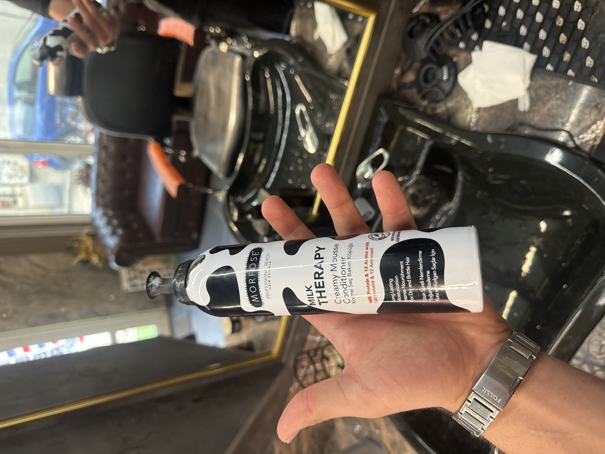 An image taken in Varna, Bulgaria, shows a person's hand holding a hair care product bottle. The image is rotated 90 degrees clockwise, with the hand and bottle appearing horizontally against a blurry background of a salon or barbershop interior. The white bottle features a distinctive black cow-print pattern and is branded "MORFOSE PROFESSIONAL REACH HAIR SCIENCE." The product is "MILK THERAPY Creamy Mousse Conditioner," described as providing "Long Lasting Moisturisation" and "Intense Nourishment for Dry and Brittle Hair." It also specifies "Milk Protein & 12 Amino acids." These descriptions are also present in Turkish ("Kremsi Saç Bakım Köpüğü," "Süt Proteini & 12 Aminoasit," "Uzun Süreli Nemlendirme," "Yoğun Besleme," "Kuru ve Kırılgan Saçlar İçin"). The hand wears a silver-toned metal watch with the "FOSSIL" brand name visible on its clasp. In the background, reflections in a gold-framed mirror show a dark, presumably leather or vinyl, barber-style chair and other indistinct elements, including a partial view of what appears to be a barber pole. The setting suggests an indoor environment, likely during the daytime, focusing on displaying the hair product.