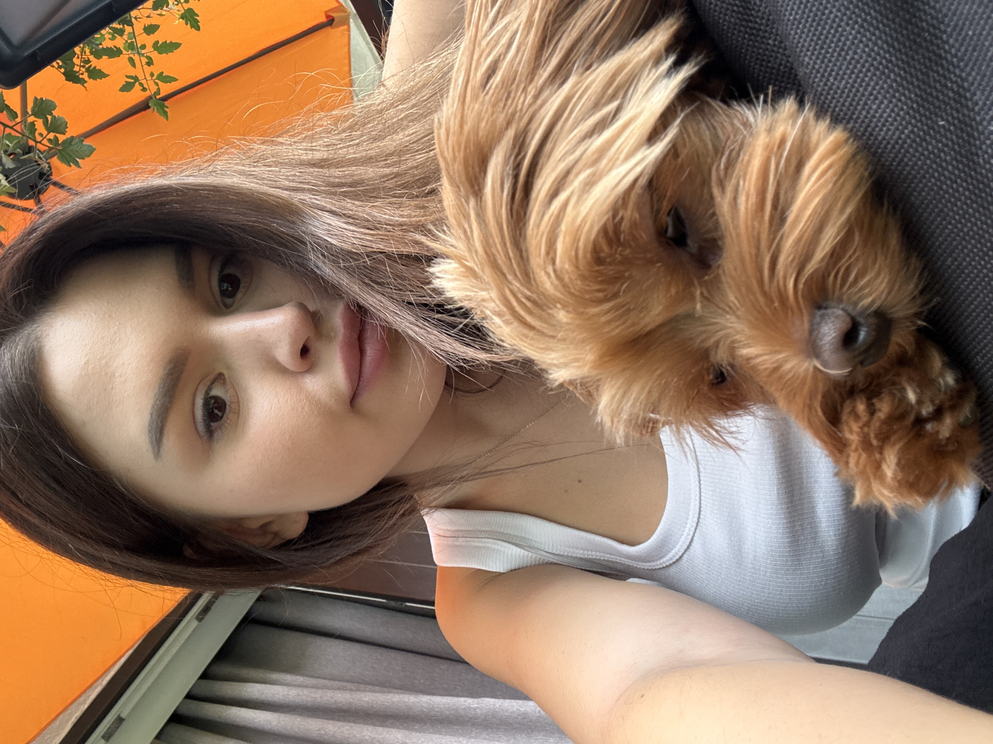 An outdoor selfie is shown, taken from a low angle. A young woman with brown hair and light skin is in the foreground, holding a small, tan-colored dog. The woman is wearing a white tank top. The dog is resting on her chest, with its face partially visible. The background is blurred, but appears to be an outdoor setting with an orange umbrella and some greenery. The overall lighting suggests it is daytime. The location is not specified as Prague, Czechia.