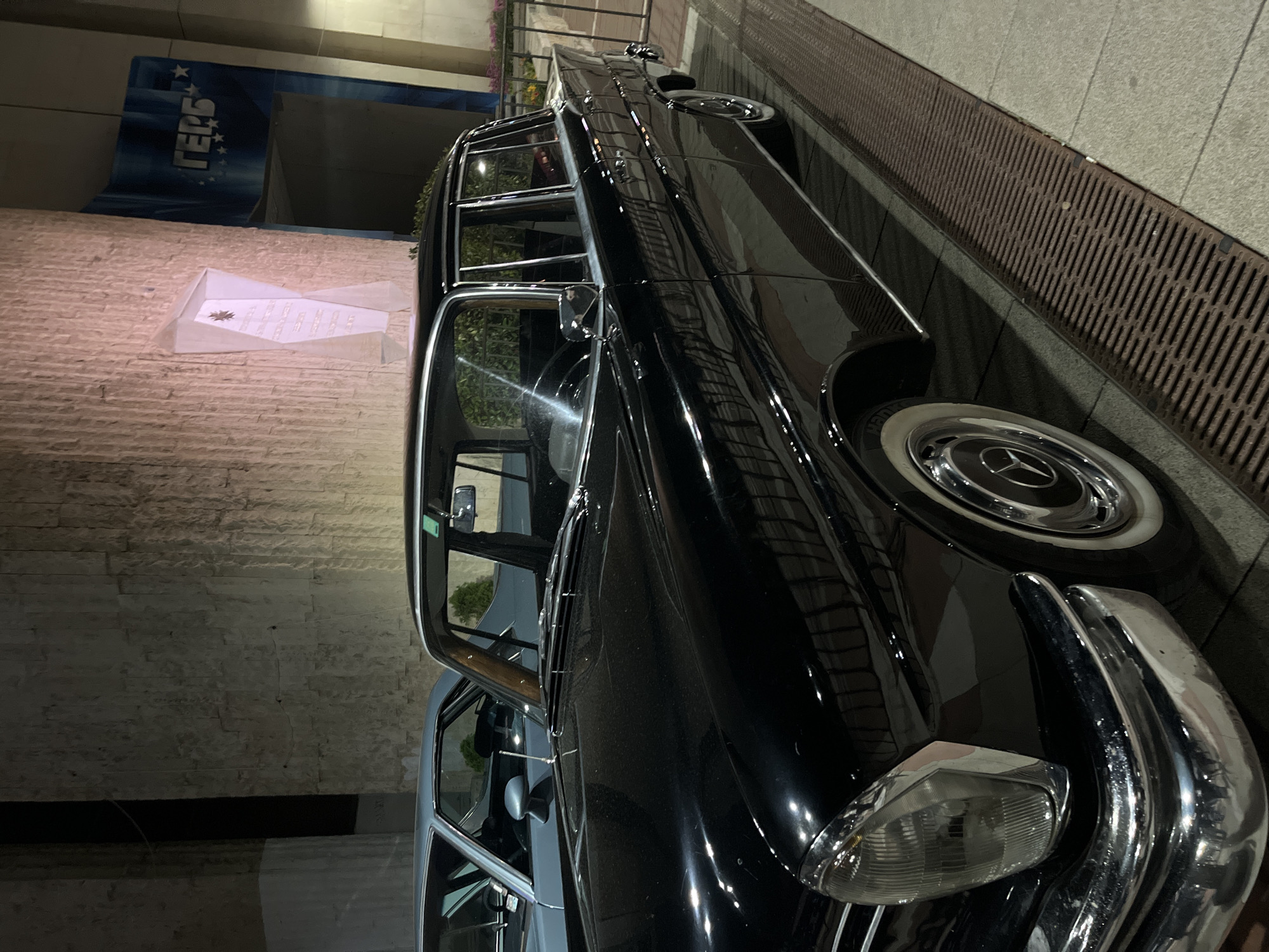 The image shows a black Mercedes-Benz limousine parked on a city street in Blagoevgrad, Bulgaria. The car is a classic model with whitewall tires and chrome accents. It is positioned alongside a sidewalk with a metal grate. Behind the car is a building with a textured brick facade and signage in Cyrillic script. The sign has the word "ГЕРБ" (GERB), a Bulgarian political party. A white paper with text or writing is placed on the wall as well. The scene is captured at night with artificial lighting illuminating the car and surrounding environment.