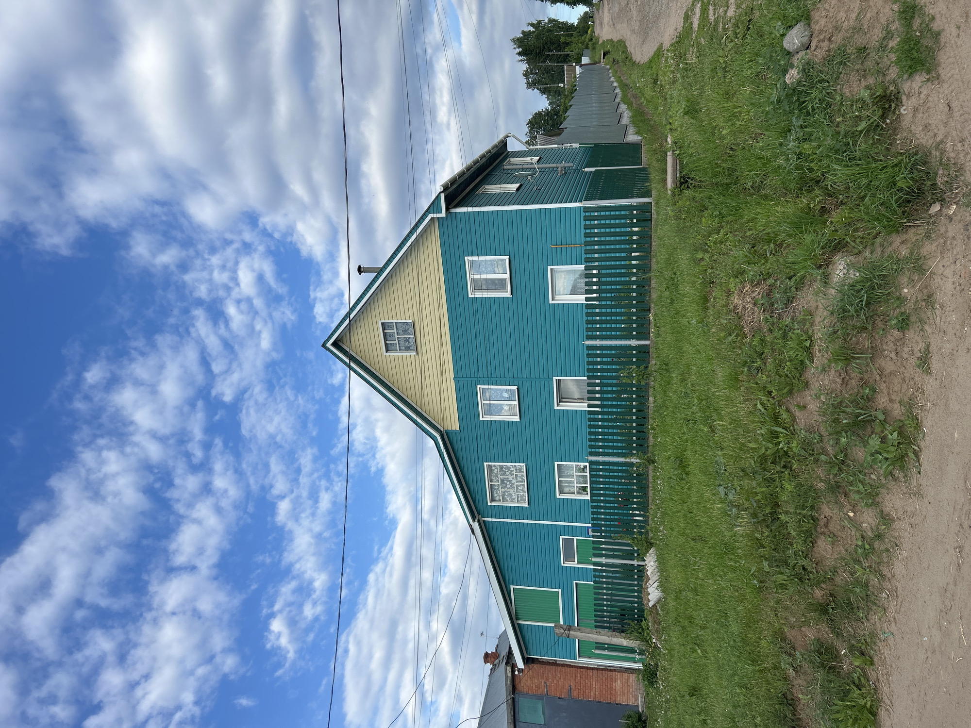 A two-story residential building, painted in vibrant teal siding with a light yellow gable end, stands under a partly cloudy blue sky. The house features multiple rectangular windows with white frames; some exhibit decorative grilles or white curtains. A dark chimney pipe protrudes from the pitched roof, which has green and white trim. On the left side of the house, large green vertical panels, resembling garage doors or shutters, are visible. White gutters and downspouts are present on the right facade.
In front of the house, a dark green picket fence encloses a grassy area. Another taller, solid green fence extends along the right side of the property, bordering a light brown dirt road that recedes into the distance and slightly uphill, flanked by trees. The foreground consists of a dirt verge leading to an unkempt grassy slope. To the left of the main house, a brick structure with a grey roof and a large dark opening is partially visible. Multiple black utility lines traverse the upper portion of the image. The scene is static, devoid of people or discernible activity, depicting a residential or semi-rural setting in Chaikovsky, Russia.