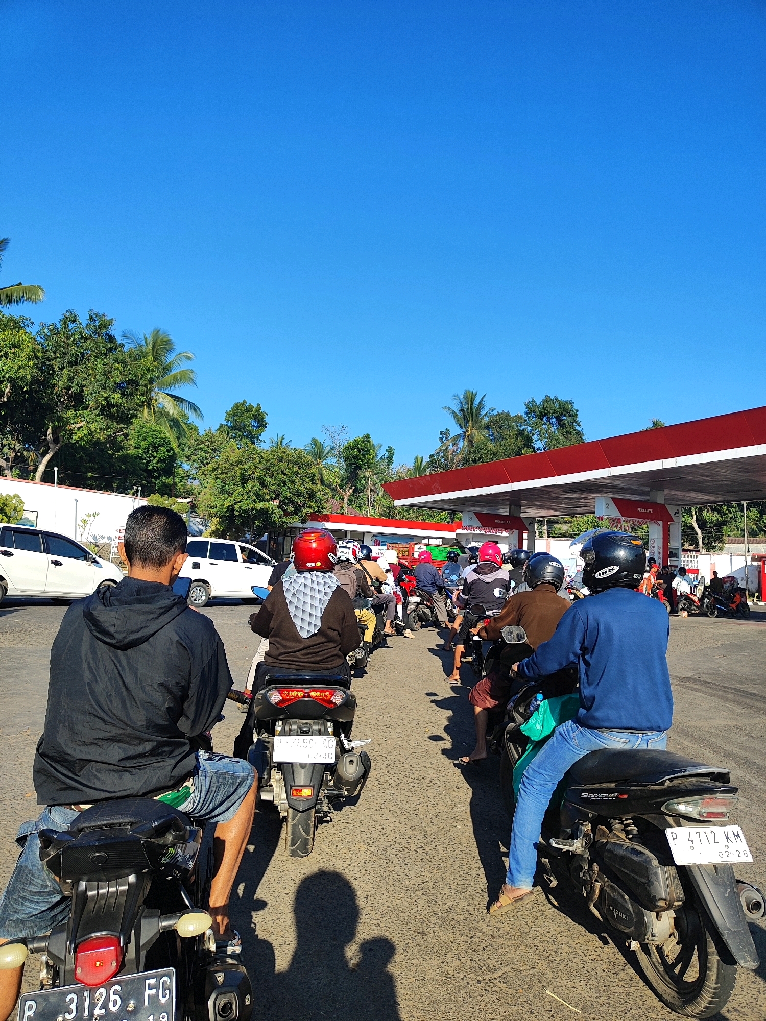 An outdoor, medium shot captures a queue of motorcycles and their riders at a gas station under a clear blue sky. The gas station features a red and white canopy extending over multiple fuel pumps. The road surface is asphalt, and trees line the background.
In the foreground, a person wearing a black hooded jacket and blue jeans sits on a motorcycle, facing away from the camera. Their motorcycle has a license plate that reads "P . 3126 . FG". To their right, another person in a blue long-sleeved shirt and blue jeans is also seated on a motorcycle, with a license plate showing "P 4712 KM 02-28".
Further down the queue, more individuals on motorcycles are visible, many wearing helmets of various colors, including red, pink, and black. Some of these riders are wearing casual clothing. In the middle ground, two white cars are parked or waiting. The overall scene depicts a typical day at a gas station, possibly experiencing a rush of customers. The presence of "P" on the license plates suggests this location is in Indonesia, and the specific format of the plates could indicate a particular region, but this cannot be definitively confirmed from the image alone without further context. Given the user's prompt to include the city of Bondowoso, Indonesia, the image is assumed to be taken there.