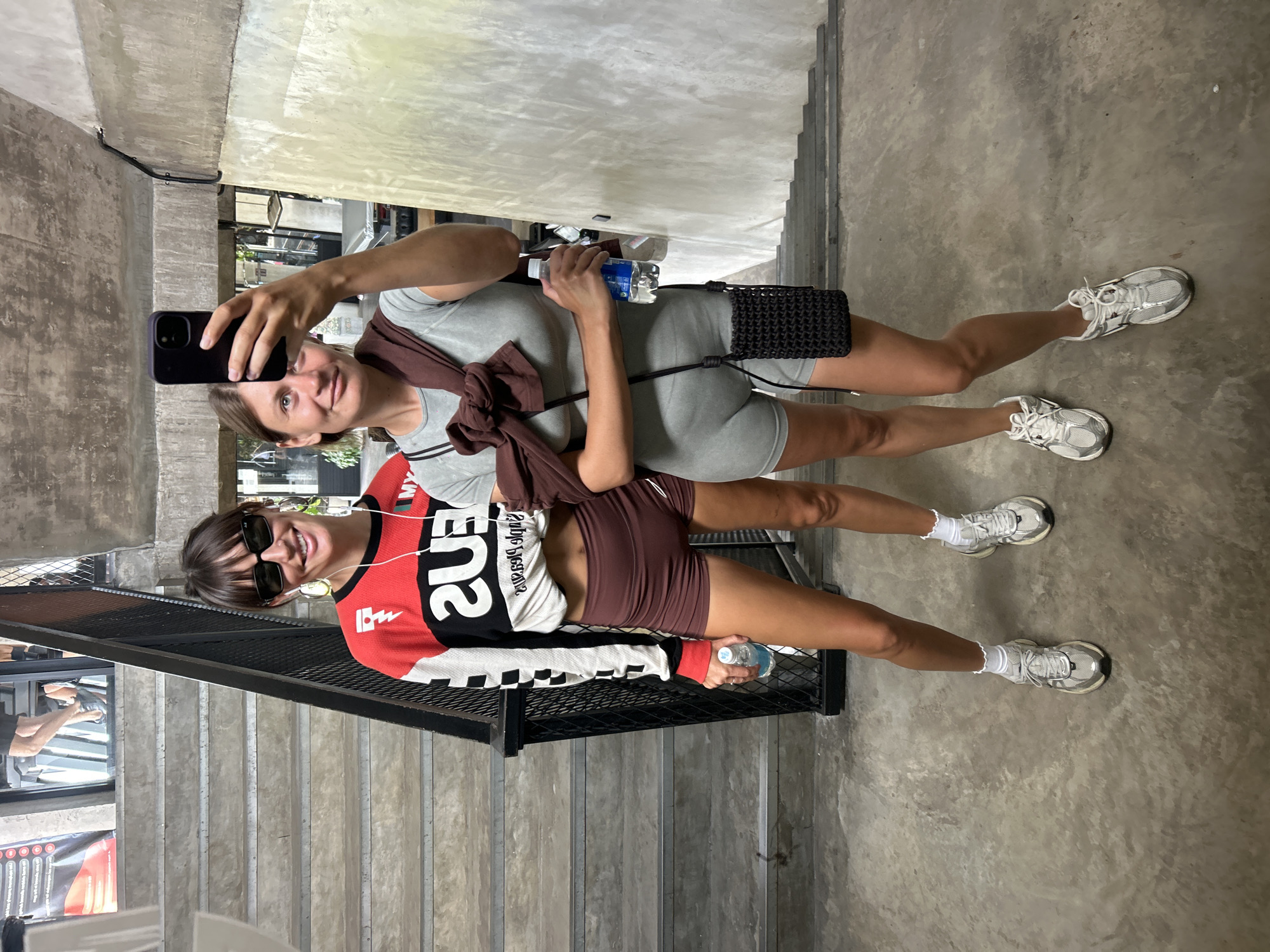 Two women are captured in a mirror selfie inside what appears to be a modern building with an industrial aesthetic, featuring concrete walls and floors, and a black metal railing with stairs visible in the background and foreground. The setting suggests a gym, studio, or retail space in Mengwi, Indonesia.
The woman taking the selfie is on the left, holding a black smartphone. She wears a grey, form-fitting athletic bodysuit with a brown long-sleeved top tied around her waist. She has fair skin, light brown hair, and is looking directly at the camera with a slight smile. Her accessories include a small black woven shoulder bag and white sneakers. She holds a clear plastic water bottle with the brand "AQUA" visible on the label.
The second woman stands slightly behind and to the right of the first, also looking at the camera with a smile. She has dark hair with bangs, is wearing black sunglasses, and has white earbuds in her ears. Her attire consists of a red, black, and white long-sleeved athletic top, with the word "SUPER" prominently displayed in white on the front. She pairs this with dark brown fitted shorts, white frilly socks, and white sneakers. She also holds a clear plastic water bottle. Both women appear to be dressed in comfortable, active wear. The overall mood is casual and friendly.
