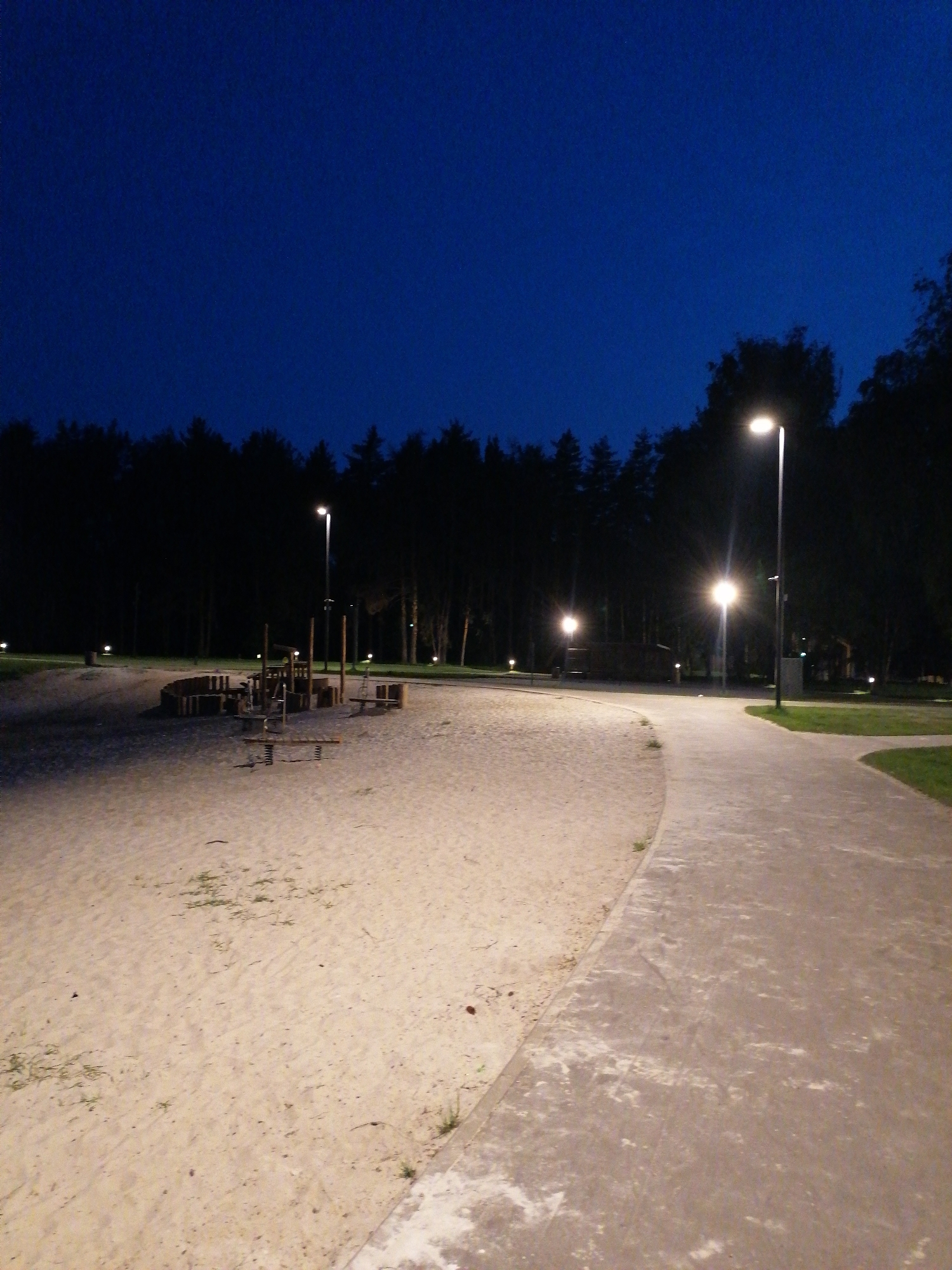 The image depicts an outdoor scene at night. It appears to be a playground area adjacent to a paved walkway. The playground is covered in sand, and there are playground structures present. Trees line the perimeter of the playground area and the walkway. Streetlights illuminate the scene. The sky is dark blue, suggesting it is late evening or night. There are no people visible in the image.