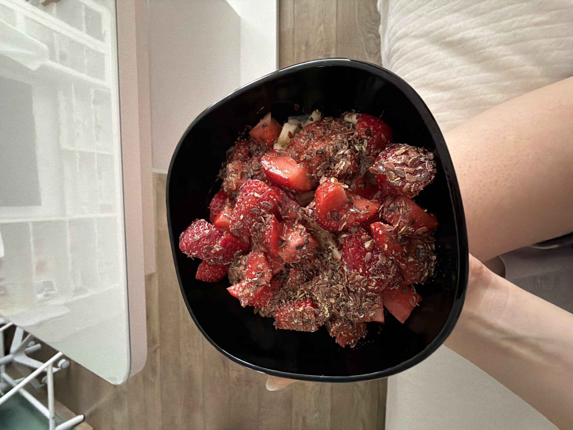 The image shows an overhead view of a person holding a black bowl filled with a healthy meal or snack. The bowl contains a substantial portion of chopped red strawberries, interspersed with lighter fruit pieces that appear to be banana. These fruits are generously topped with a mixture of dark brown flakes and lighter seeds, likely flax seeds and possibly cacao nibs or granola.
The person holding the bowl is partially visible, with their forearms and hands supporting it. A portion of their bare leg is visible on the right side of the frame, resting on a white, textured fabric, which could be a blanket or bedding.
The setting appears to be indoors, likely a home in Helmond, Netherlands. To the left of the bowl, a tall, reflective surface, possibly a refrigerator or a cabinet door, shows a blurred reflection of what seems to be windowpanes or interior shelving. Below this, a white metal grid structure, resembling a rack or shelf, is partially visible. The background also features a wooden surface, suggesting either a table, floor, or wall. The scene evokes a casual and relaxed atmosphere, implying a meal being enjoyed in comfort, possibly during breakfast or a break. No text is visible in the image.