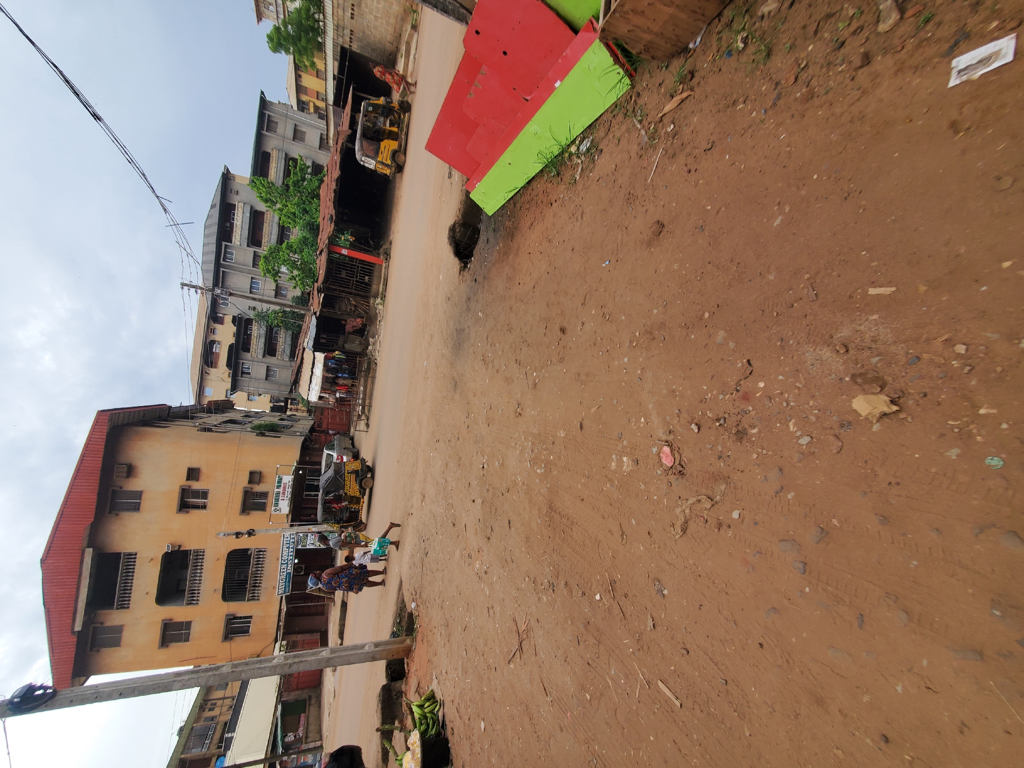 The scene is located in Onitsha, Nigeria. A dirt road is flanked by multi-story buildings, some with balconies. The road has a large pothole near a stack of red and green painted materials. There are utility poles and wires overhead. People are present, including a woman and child walking by a building with commercial signs ("MAYGREEN COMPUTER INSTITUTE" visible). There's also a yellow tricycle vehicle ("keke") in the scene. Some produce, possibly plantains, are on the ground near a pole. The sky is overcast.