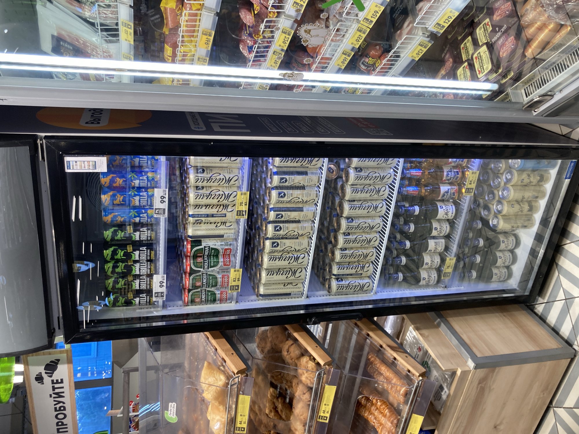 This is an eye-level, medium shot of a commercial refrigerator in a grocery store, likely in Moscow, Russia, given the Cyrillic text on product labels and signage. The refrigerator door is made of glass, allowing a clear view of its contents.
Inside the refrigerator, the shelves are stocked with various beverages, primarily canned drinks and bottled water. The top shelves are filled with cans featuring designs with green and blue characters, suggesting energy drinks or similar products. Below these are rows of cans with a distinct white and gold label, some appearing to be beer. Further down are rows of bottled beverages.
To the left of the refrigerator, display shelves are filled with baked goods, including pastries and bread products. The signage above these shelves and on the refrigerator displays Cyrillic text, which translates to "Try it" and other promotional messages.
The refrigerator is situated next to shelves stocked with meat products, including sausages and packaged cured meats. These shelves are also labeled with Cyrillic text and price tags. The overall environment suggests a well-stocked retail space. The lighting is bright and commercial, with the refrigerator's internal lighting illuminating the products. The floor is tiled with a diagonal striped pattern. No people are visible in the image.