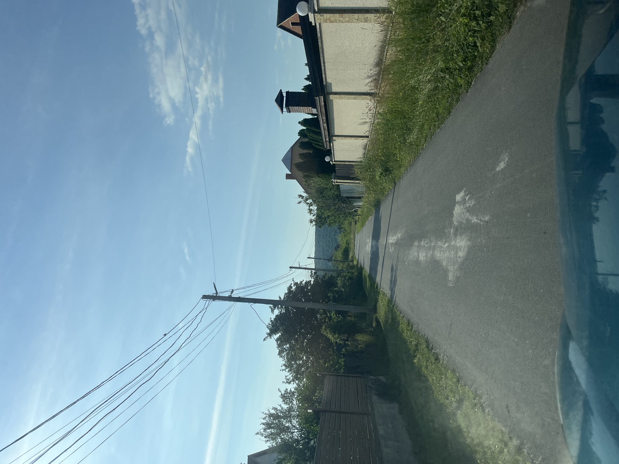 The image depicts a view from a vehicle driving down an asphalt road in Onokivtsi, Ukraine. The road is lined with fences and vegetation on both sides. On the left, a wooden fence is visible, with trees and foliage behind it. On the right, a tall, beige stucco wall with stone accents runs along the road, topped by decorative spherical lights. Behind this wall, houses with distinct architectural features, including a house with a pyramidal roof and another with a brick facade and chimney, are partially visible. The sky above is a clear blue with scattered white clouds. Power lines stretch across the sky, supported by utility poles. The overall scene suggests a residential area on the outskirts of the town, characterized by private homes and a rural road environment.