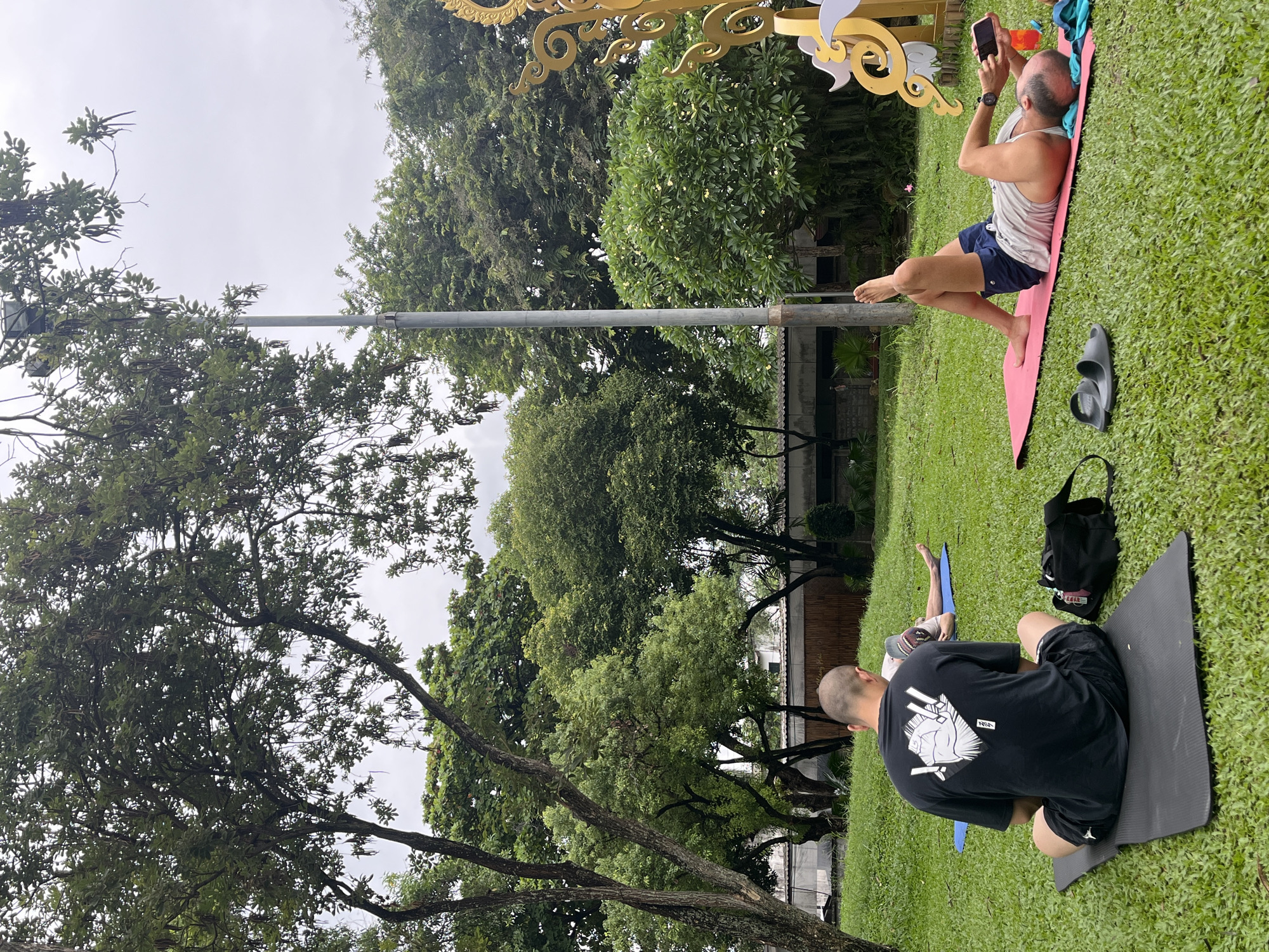 In Chiang Mai City Municipality, Thailand, an outdoor yoga class is taking place in a grassy park. Three individuals are visible, each on their own yoga mat. In the foreground, a bald man wearing a black t-shirt with a graphic design and black shorts sits cross-legged on a black yoga mat. Next to him is a black gym bag and a pair of black sandals. Further back, another man lies on his back on a pink yoga mat, wearing a grey tank top and blue shorts. He is holding a smartphone in his right hand and has a water bottle nearby. A third individual, partially visible, is lying on their side on a blue yoga mat in the background. The setting is lush and green, with mature trees and foliage surrounding the area. A lamppost stands in the middle ground, and a decorative golden structure is visible on the right edge of the frame. The sky is overcast.
