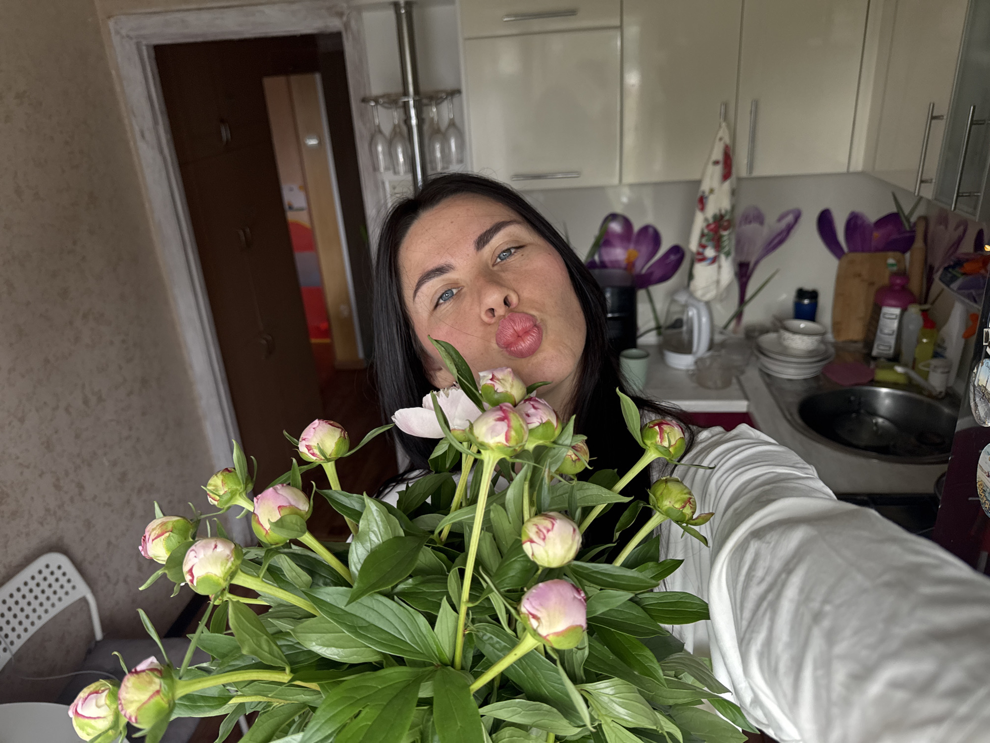 A woman with dark hair and blue eyes is holding a bouquet of peonies and pouting her lips. She is wearing a white shirt. She is in a kitchen, with kitchen cabinets, a sink, and a stovetop visible in the background. There is a doorway in the background that leads to another room. The walls of the kitchen are decorated with purple flower decals. The lighting is dim, and the overall scene is casual and domestic. The presence of kitchen appliances and cabinets suggests the image was taken in a residential kitchen.