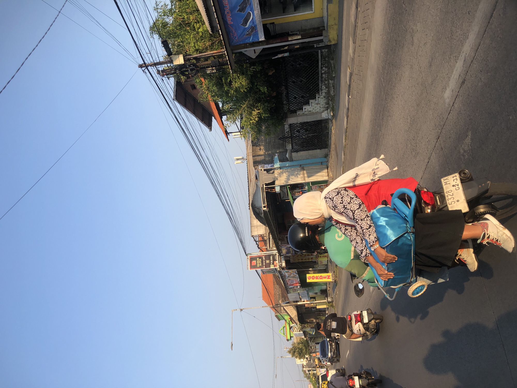 In the city of Boro, Indonesia, a couple is riding a motorcycle. The man, wearing a black helmet and a green shirt, is driving. The woman, wearing a white hijab and a patterned top, is sitting behind him, with her arms around his waist. She appears to be kissing him on the cheek. They are both carrying a blue bag with wheels. The motorcycle is in motion on a paved road, with other vehicles and buildings visible in the background. The sun is low in the sky, casting long shadows on the road, indicating it is either early morning or late afternoon. The environment appears to be a street in a bustling urban or suburban area.