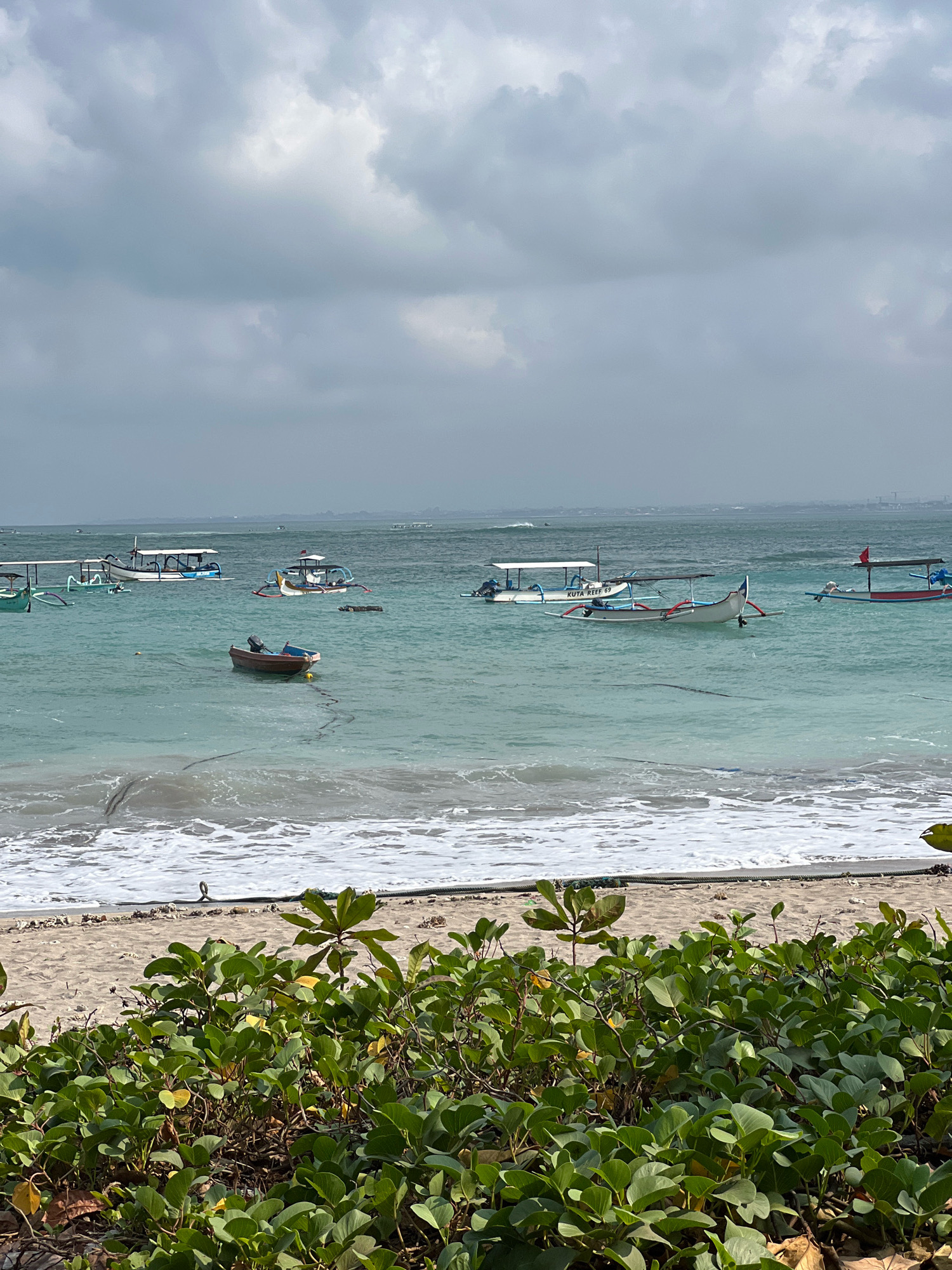 The image shows a tranquil beach scene in Kuta, Indonesia. The foreground is dominated by lush green vegetation, casting shadows on the sandy beach. The ocean is a calm turquoise, with gentle waves lapping at the shore. Several traditional Indonesian fishing boats, known as jukungs, are moored in the shallow water, some with outriggers. The sky is overcast with soft grey clouds, creating a diffused light. In the distance, a faint outline of buildings can be seen on the horizon. The overall atmosphere is serene and peaceful, characteristic of a relaxed coastal environment.