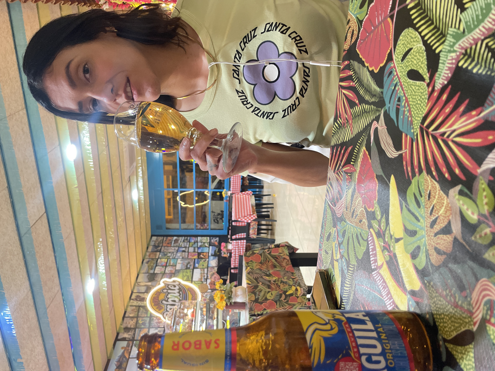 A woman in a light green "Santa Cruz" t-shirt is holding a glass of beer. The table is covered in a patterned, tropical-leaf-motif tablecloth. A bottle of "Aguila" beer is on the table. The background shows a restaurant interior, with a colorful wall featuring horizontal stripes and what appears to be photo collages. A neon sign that says "Gabriel" is visible, and through the glass door, there is a view of more tables and chairs. The location is the Gabriel Restaurant in L'Hospitalet De Llobregat, Spain.