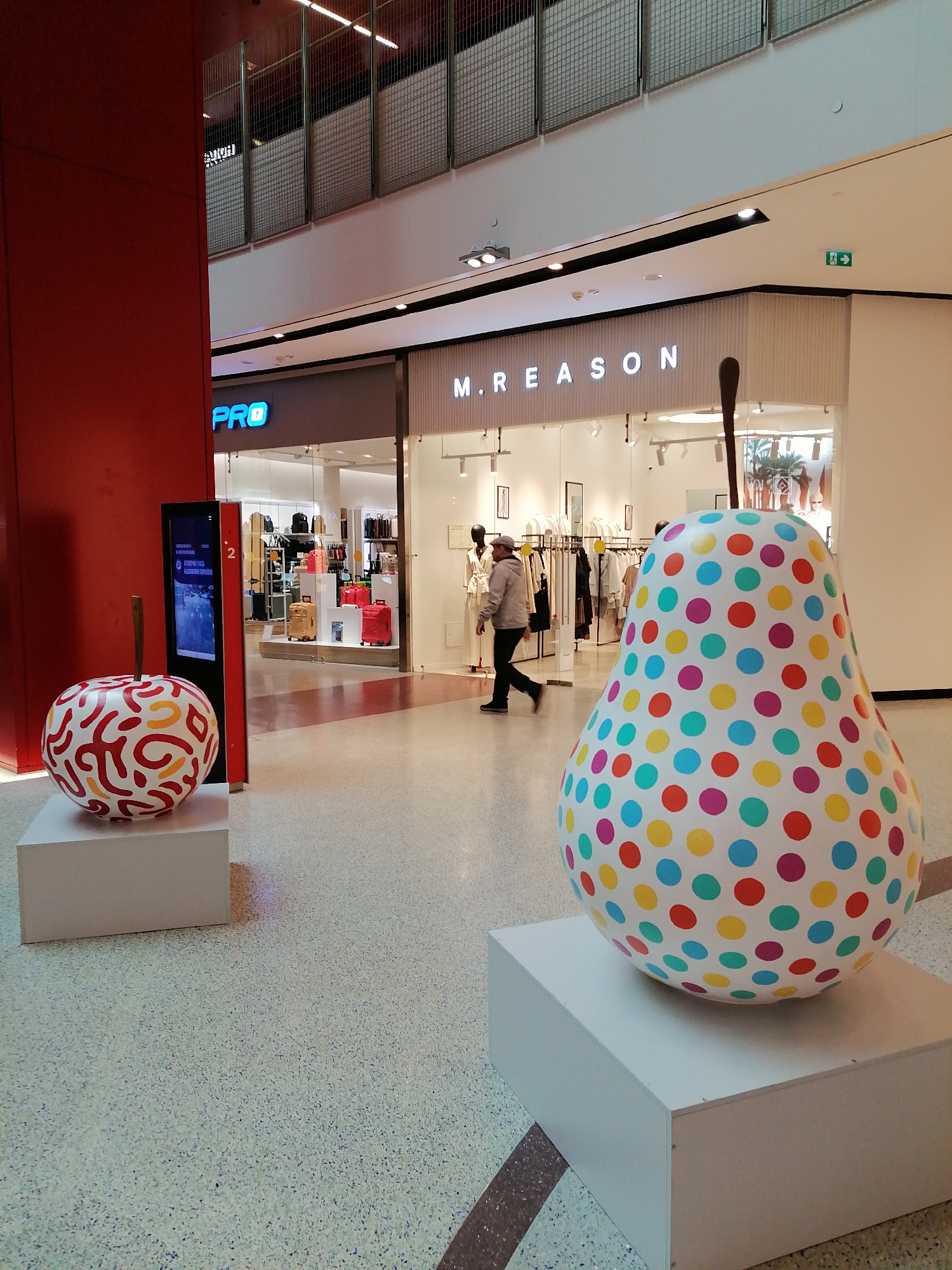 This image shows a brightly lit interior of a mall, possibly in Moscow, Russia. Two large, decorative fruit sculptures, an apple and a pear, are placed on white pedestals in the middle of the walkway. The apple has a white base with colorful, swirly designs in red and yellow. The pear is white with evenly spaced dots of various colors including blue, green, yellow, red, and purple.
In the background, there are two storefronts: one for "PRO" with clothing and luggage on display, and another for "M.REASON," a clothing store with mannequins and clothes racks visible through the glass windows. A person wearing a jacket and a cap is walking in front of the "M.REASON" store. An electronic display stands between the apple sculpture and the PRO store. The floor is a light-colored terrazzo. An exit sign is visible near the ceiling above M.REASON. Metal railings are visible in the upper background, near the ceiling.