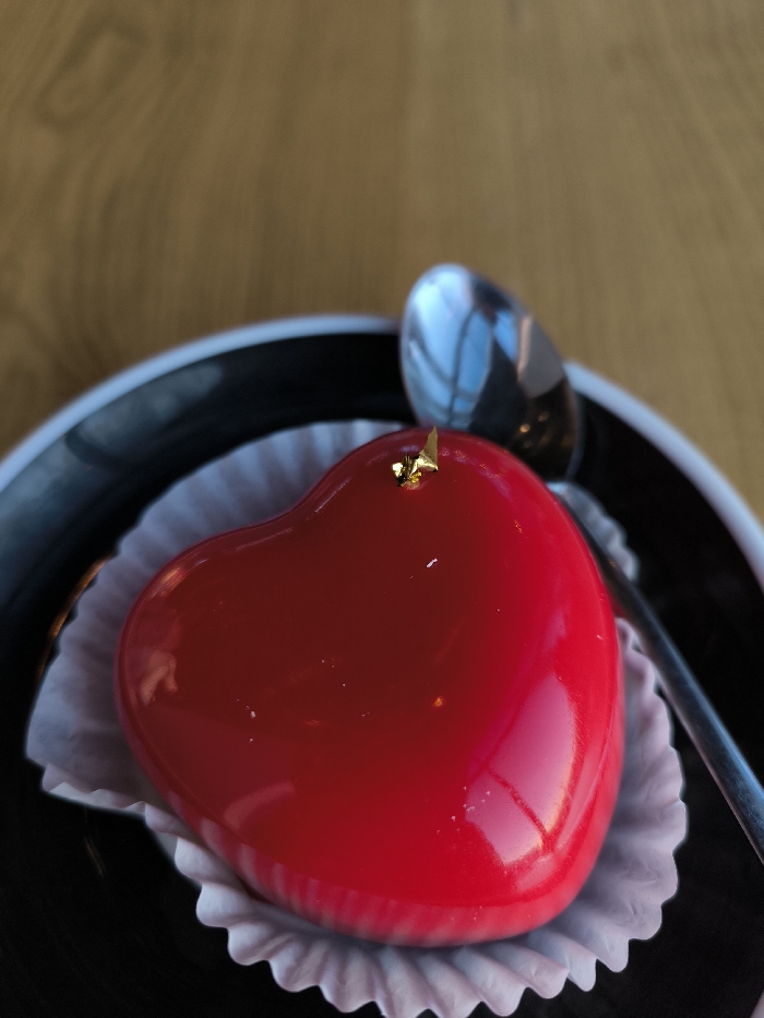 A heart-shaped dessert with a glossy red glaze is centrally positioned on a black plate with a white rim, set in the city of Vinnytsia, Ukraine. The dessert is contained within a white, fluted paper cup liner. A small, irregular piece of gold leaf is adhered to the top surface of the red glaze. A silver spoon with a reflective bowl rests on the plate to the right of the dessert, partially visible. The background consists of a blurred light brown wooden surface, displaying discernible grain patterns. No individuals or active interactions are depicted.