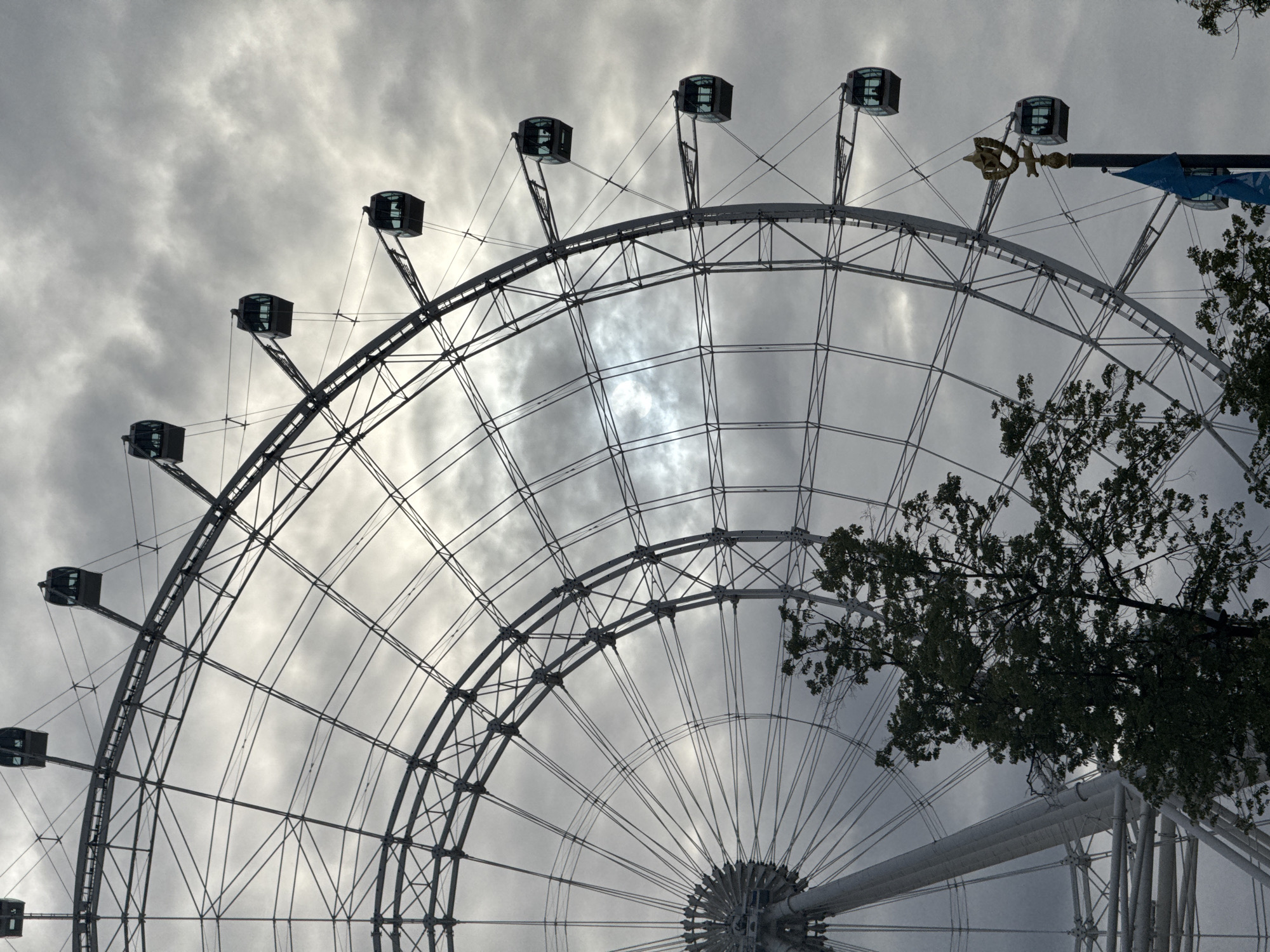 The image shows a partial view of a large Ferris wheel against a cloudy sky. The wheel is constructed of metal beams and has enclosed passenger cabins. Some trees are visible on the right side of the frame. The location is Moscow, Russia.