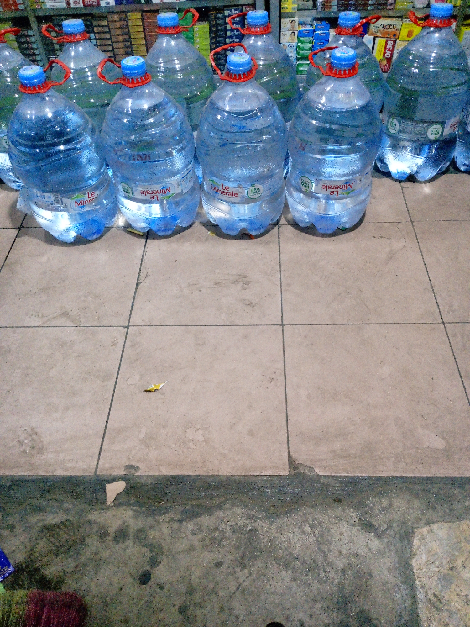 The image shows a row of large plastic water bottles with blue caps and red handles, arranged on a tiled floor. The bottles are filled with clear liquid and have labels that say "Le Minerale." In the background, shelves stocked with various products, likely in a store, are visible. The floor tiles are beige and appear to be laid in a grid pattern. The lower portion of the image shows a concrete floor with some debris. A broom with colorful bristles is partially visible in the bottom left corner. There are no people visible in the scene. The setting appears to be an indoor retail environment.