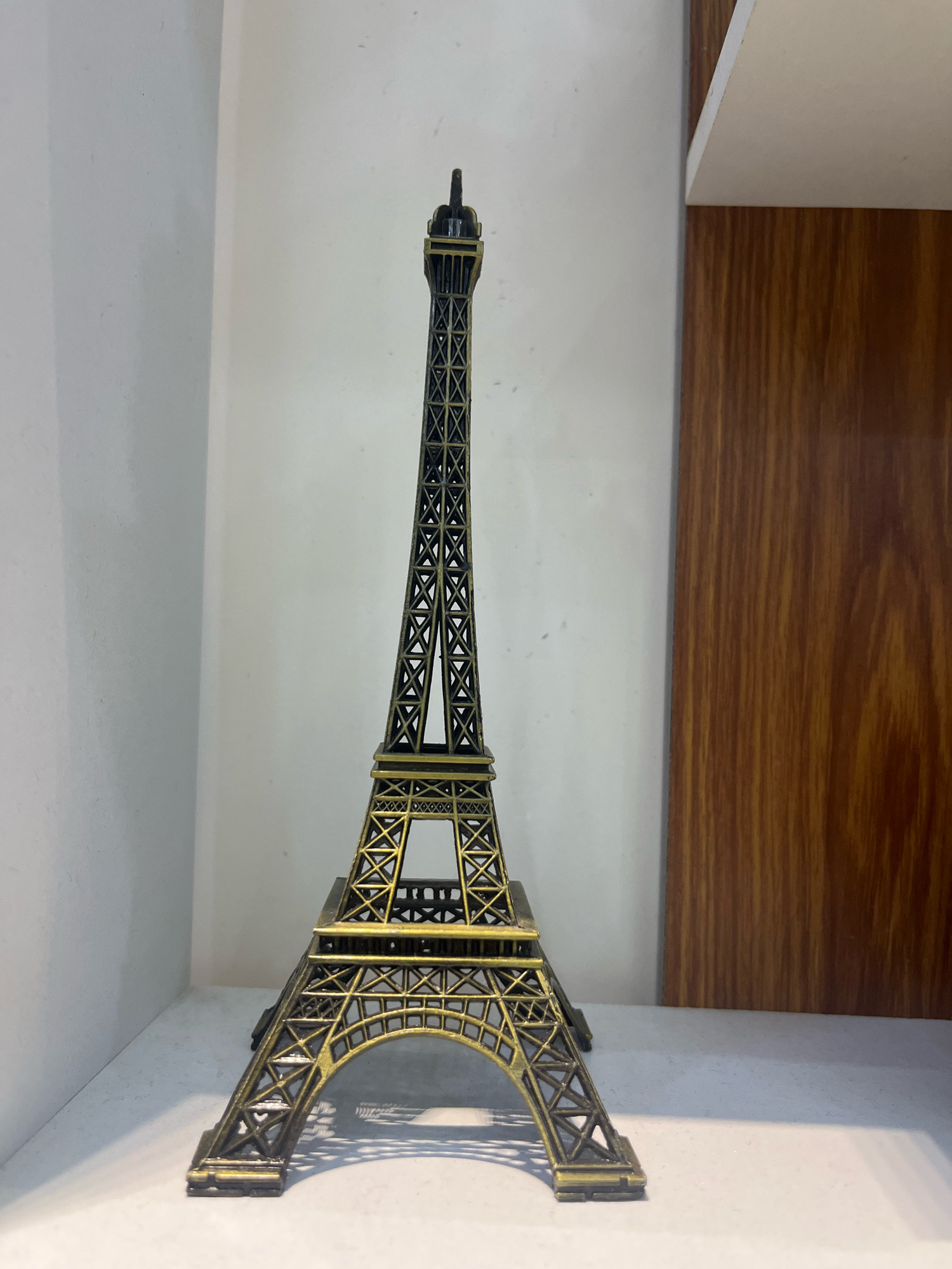 A miniature Eiffel Tower replica is positioned on a white surface. The replica is made of dark metal with golden accents and features intricate lattice work characteristic of the actual monument. To the left of the replica, a plain white wall serves as a backdrop. To the right, a vertical wooden panel with a prominent grain pattern is visible. The lighting in the scene creates soft shadows on the white surface beneath the tower. There is no indication of the city of Firozpur, India, within the image.