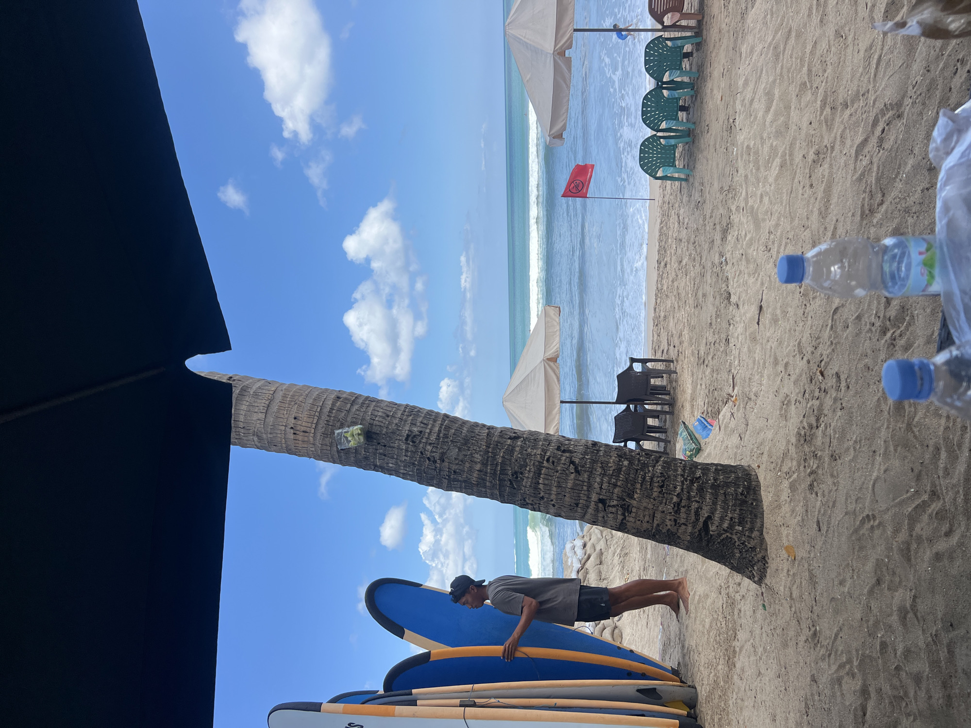 The image depicts a beach scene in Kuta, Indonesia, on a sunny day with a blue sky and scattered clouds. In the foreground, a black umbrella casts a shadow over part of the sandy beach. To the left, a man wearing a grey t-shirt and black shorts stands next to several surfboards. A tall palm tree stands prominently in the middle ground, with a small container attached to its trunk. Further back, beach umbrellas and chairs are set up on the sand. The ocean is visible with gentle waves, and a red flag is planted in the sand near the water's edge. Bottles of water and some plastic bags are present on a surface in the immediate foreground.