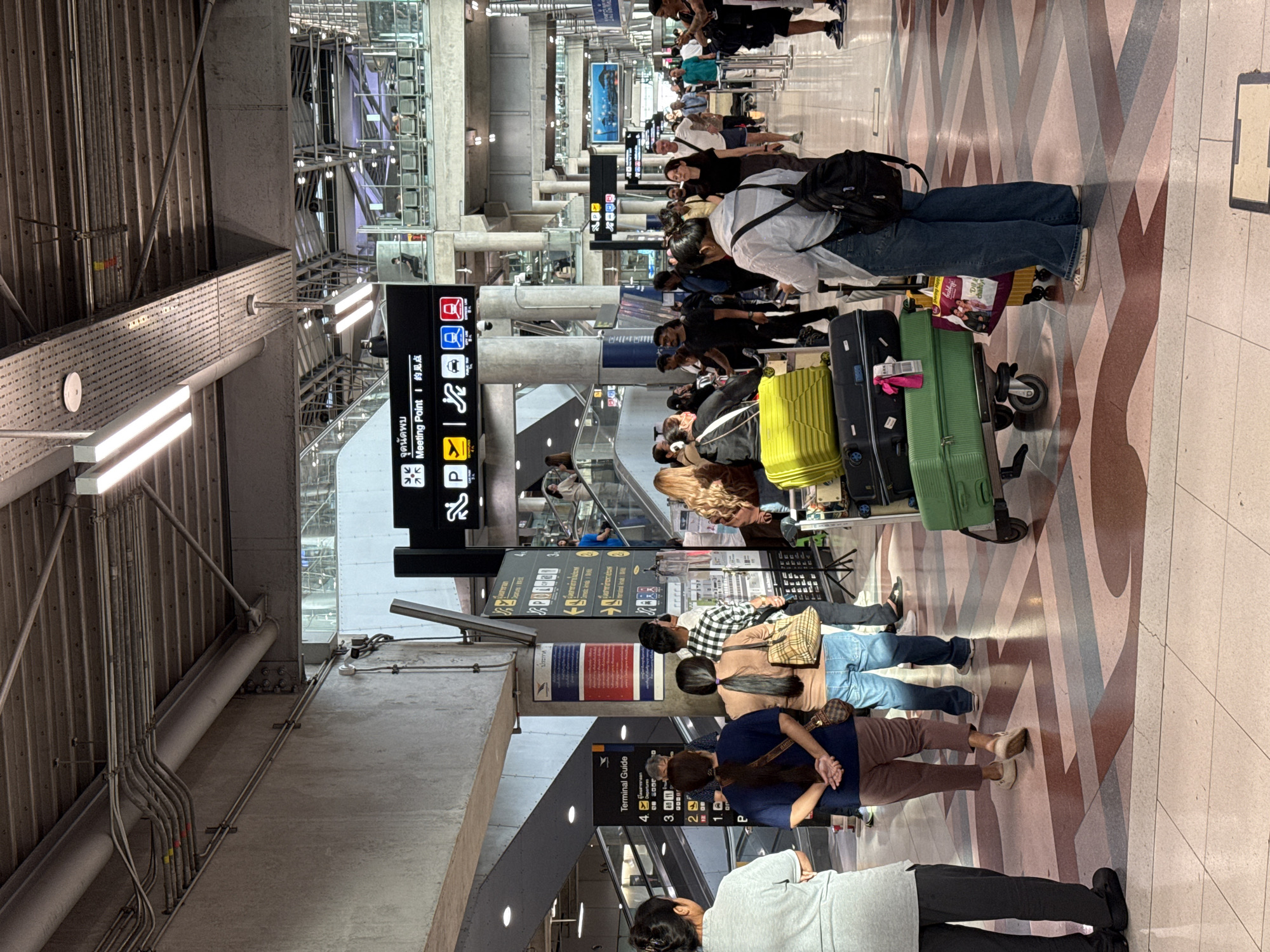 The image depicts the interior of a busy airport terminal in Nong Prue Subdistrict, Thailand. A large group of people are navigating the concourse, some with luggage. The floor is a patterned tile mosaic in shades of pink, brown, and beige. Overhead, industrial lighting fixtures and exposed conduits are visible within the ceiling structure.
Several directional signs are present. A prominent black sign with white text displays "Meeting Point" in English and Thai script, accompanied by symbols for a meeting point, a plane, a taxi, and a public transportation bus. Another sign, "Terminal Guide," lists numbered levels.
People are seen standing, walking, and pushing luggage carts. One cart is laden with several suitcases, including a bright yellow one and a green one. Individuals are dressed in casual attire. The overall atmosphere suggests travel and transit. The architectural style of the terminal appears modern and expansive.
