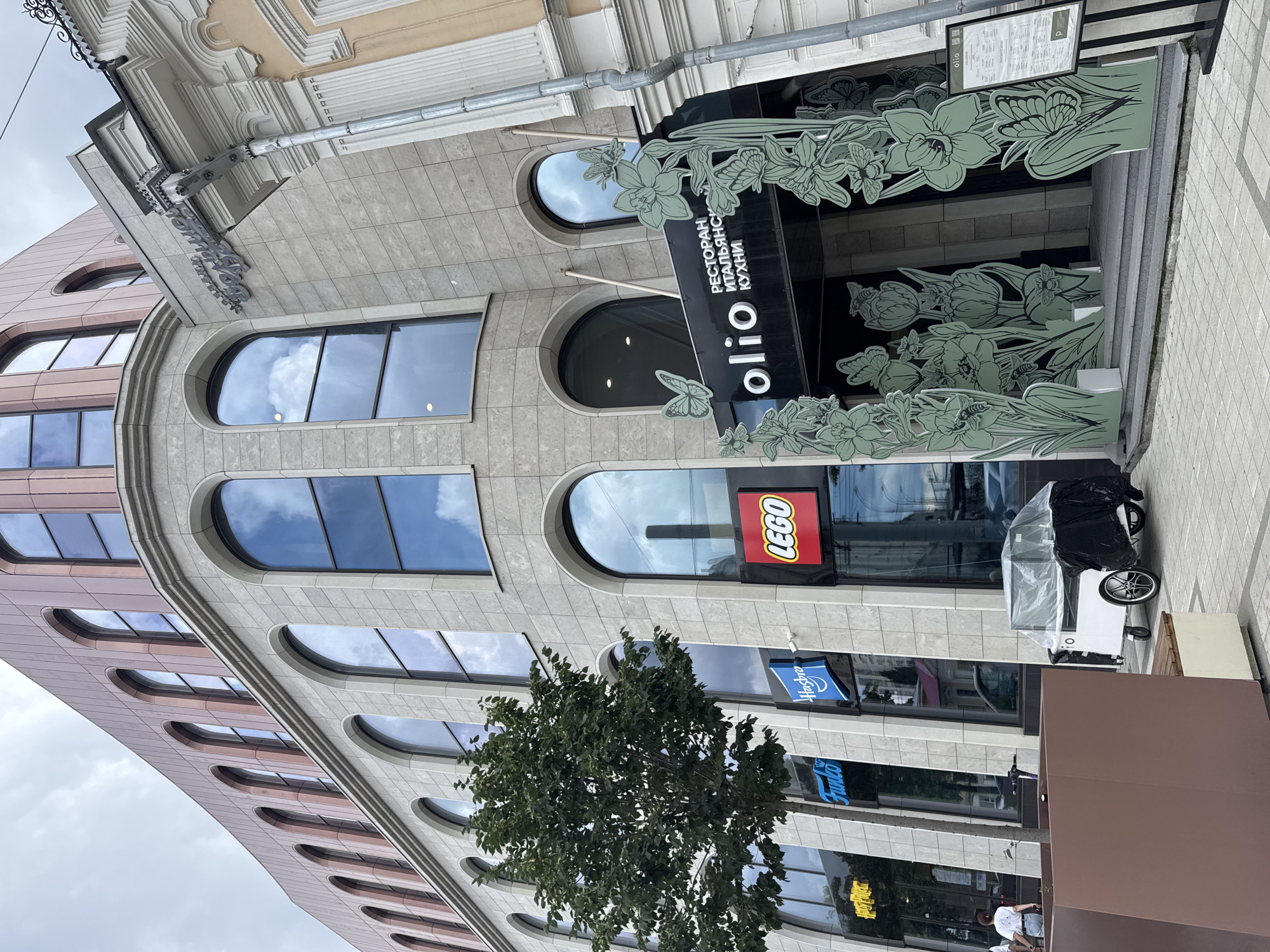 The image shows a building in Yekaterinburg, Russia, with a facade of light-colored stone and arched windows. Several storefronts are visible on the ground floor, including "Harry Potter," "Funko," "Hasbro," and "LEGO." Above the entrance to "Olio," a restaurant with Italian cuisine, there are decorative green cutouts of flowers and butterflies. A small, white cart covered with plastic sheeting is parked on the sidewalk in front of the building, near a brown planter. The sky is overcast. A person in a white shirt and shorts is walking away from the camera on the left side of the image. Another person is partially visible behind the planter.