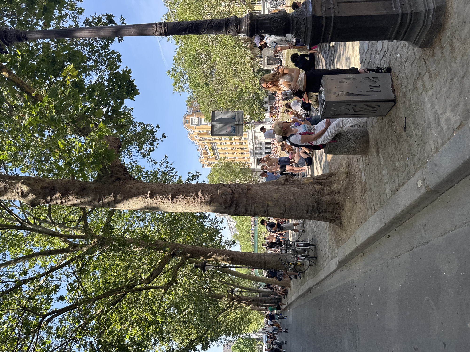 The image shows a sunny day with a clear blue sky in Paris, France, likely along a canal or riverbank, given the presence of people relaxing by the water. The scene is lined with mature trees providing shade, with a paved walkway and a street. A diverse group of people are present, some walking or cycling on the street, while others are sitting or standing on the walkway, socializing or picnicking. There are bicycles parked along the side of the path and a decorative lamppost in the foreground. Buildings with distinct architecture are visible in the background, suggesting an urban setting. The overall atmosphere appears relaxed and leisurely, with many individuals enjoying the outdoor environment.