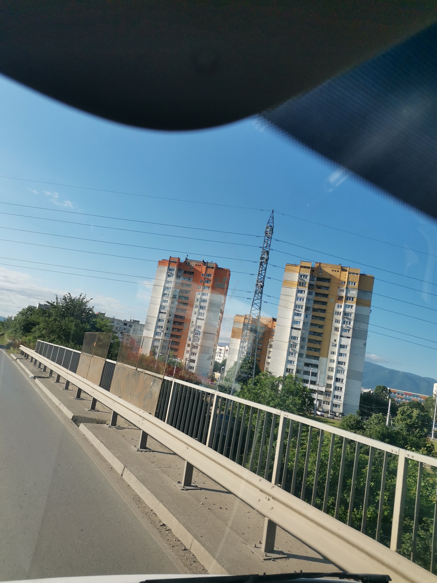 The image shows a view from a moving vehicle, likely a car, looking forward and slightly to the right. In the foreground, a concrete barrier with a metal guardrail runs along the side of a road. Beyond the barrier, there are several tall residential buildings, two of which are prominent in the midground. One building has an orange and white facade, while the other has a yellow and white facade. A large metal pylon with electrical wires stretches upwards between these two buildings. Lush green trees and foliage are visible in the midground and background, with more buildings and a mountain range in the far distance. The sky is a clear blue with a few wispy clouds. The scene appears to be in an urban or suburban area, possibly on a bridge or elevated road, given the perspective. The architectural style of the buildings suggests a post-Soviet era construction, common in cities like Sofia, Bulgaria. This scene is consistent with views found in Sofia's residential districts like Mladost or Lyulin, areas known for their high-rise buildings and proximity to infrastructure like power lines and major roads.