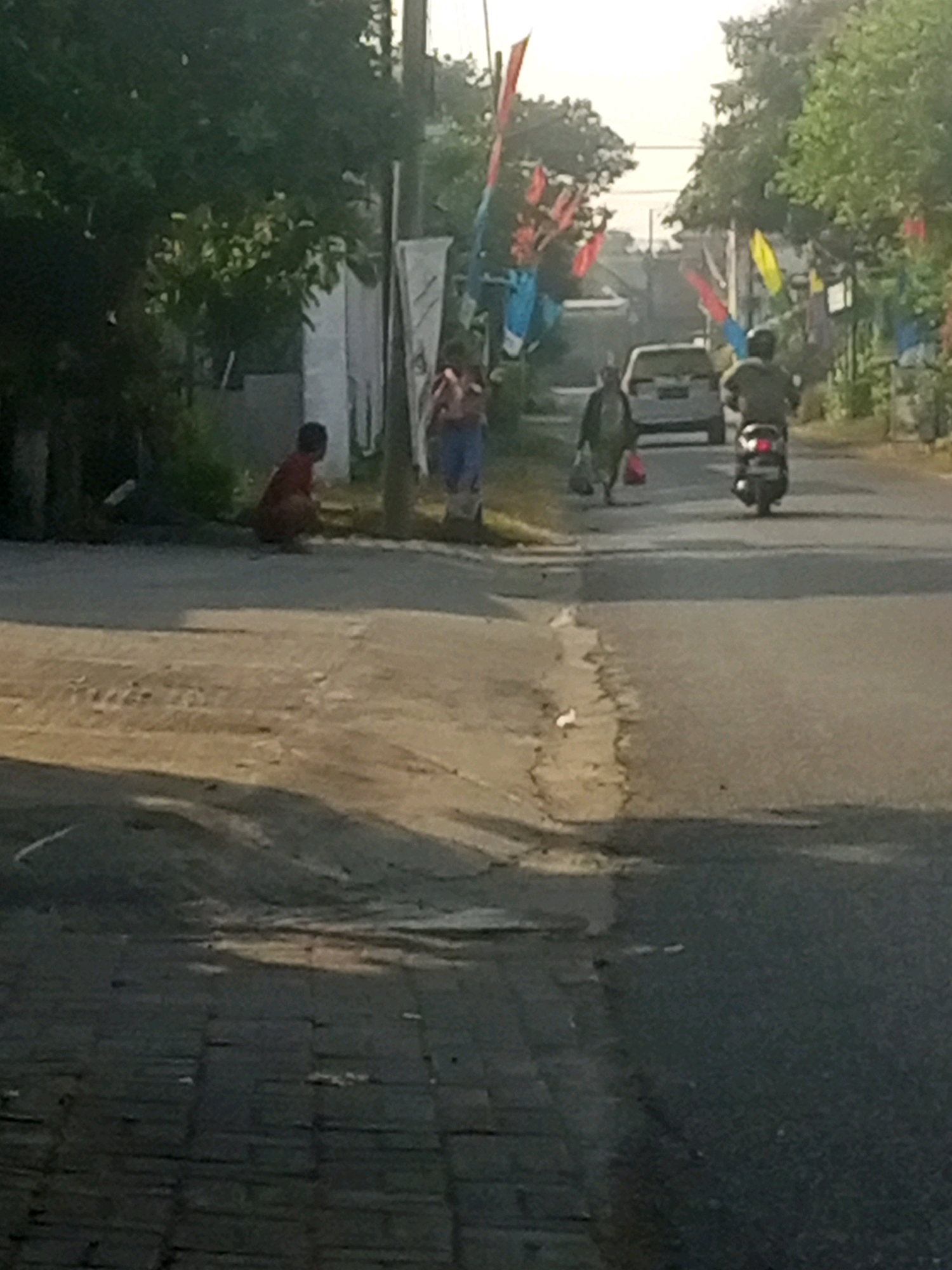 A street scene in Gurah, Indonesia, depicts a young child in a red shirt squatting on the side of the road. Further down the road, a person walks away from the camera carrying two plastic bags. A white car is visible in the middle ground, and a person on a scooter rides away from the camera in the right lane. The road is lined with trees and decorated with colorful flags. The pavement on the left side of the road is made of interlocking blocks.