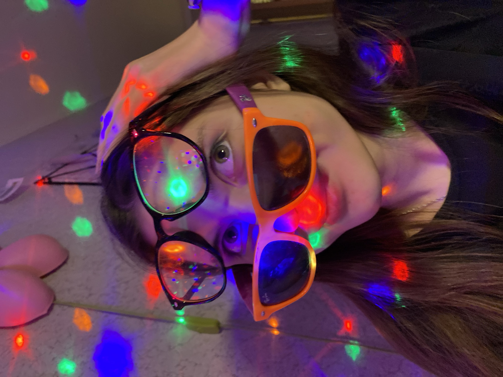 A person lies on a surface illuminated by colorful lights, possibly from a disco ball or string lights. The individual is wearing two pairs of glasses: one pair has black frames and clear lenses, and the other pair has orange frames with darker lenses. The person's hair is dark and shoulder-length. Two pink, dome-shaped objects are visible in the lower left corner of the image. The scene appears to be indoors.
It is not possible to verify the location in the city of Votkinsk, Russia from the provided image.