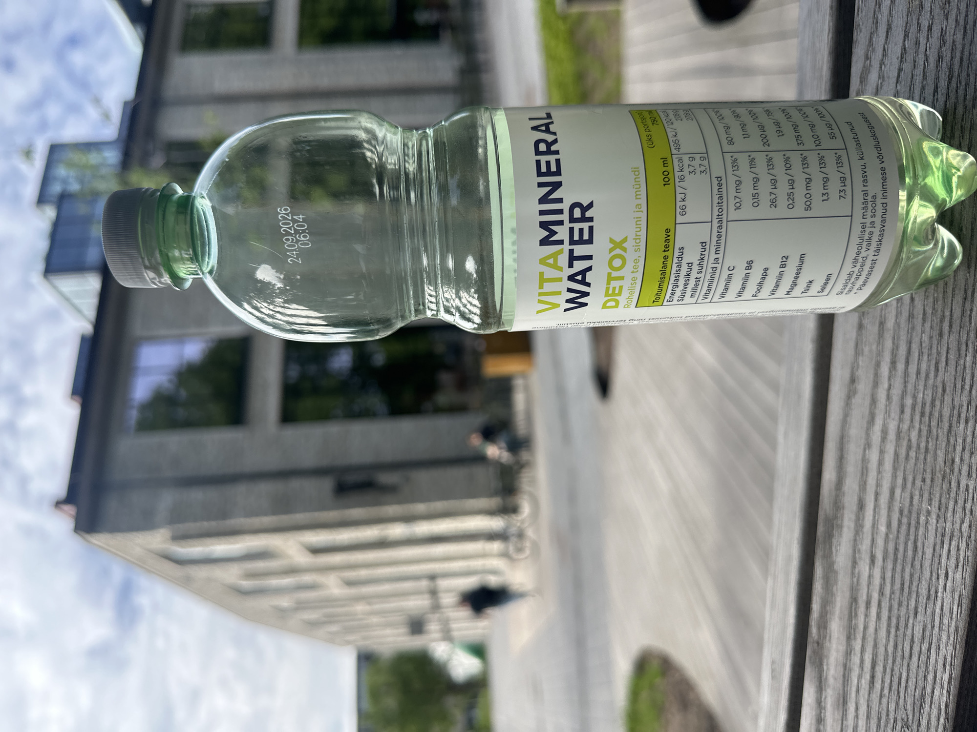 An outdoor close-up shot features a bottle of "VITAMIN MINERAL WATER DETOX" positioned on a wooden surface, likely a table or bench. The bottle's label is visible, displaying nutritional information in Estonian, and the bottle itself has a pale green tint. In the background, a modern, grey brick building with large windows stands under a cloudy sky. The building's architectural style suggests it could be a contemporary public or commercial structure, possibly a university building or office complex. A blurred street scene unfolds in front of the building, with a few individuals and a bicycle visible. One person is walking away from the camera, and another is seated on a bench. The overall setting appears to be an urban environment in Tallinn, Estonia, possibly a park or a campus area adjacent to the building.