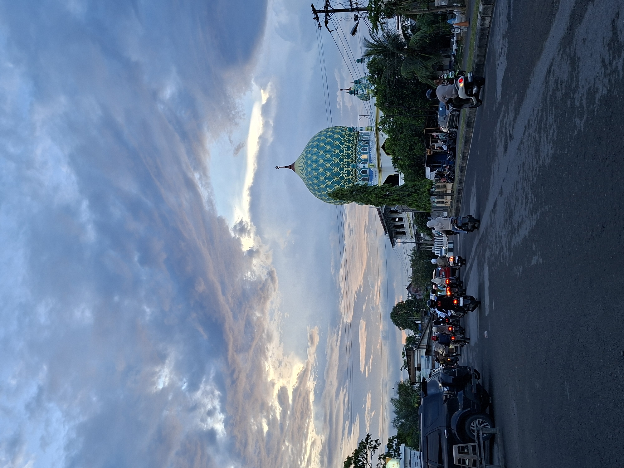 The image captures a street scene in Lhokseumawe, Indonesia, dominated by a mosque with a distinctive blue and gold patterned dome. The sky features dynamic cloud formations with hints of sunset colors. Traffic, primarily motorbikes, is visible on the asphalt road. Several individuals are riding or standing near the parked motorbikes. Buildings and vegetation line the street, including trees and power lines. A black SUV is parked in the foreground.