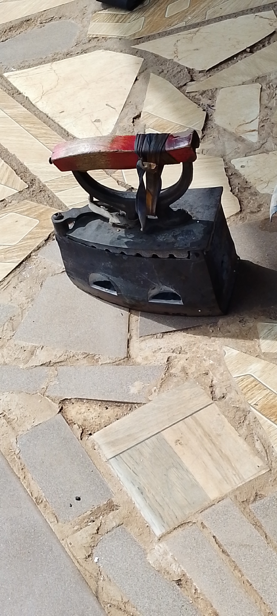 An antique, black, cast iron clothes iron with a red wooden handle is situated on a patterned ground made of irregular, flat stones. The iron is positioned on its side, revealing two rectangular openings on its body. The handle is secured by dark, wrapped material. The ground beneath the iron is a mosaic of light brown, beige, and grey stones of varying shapes and sizes, with visible dirt and debris in the gaps between them. The lighting suggests an outdoor setting, possibly a courtyard or walkway. The overall scene appears static, with no visible activity. The location is specified as Kachia, Nigeria.