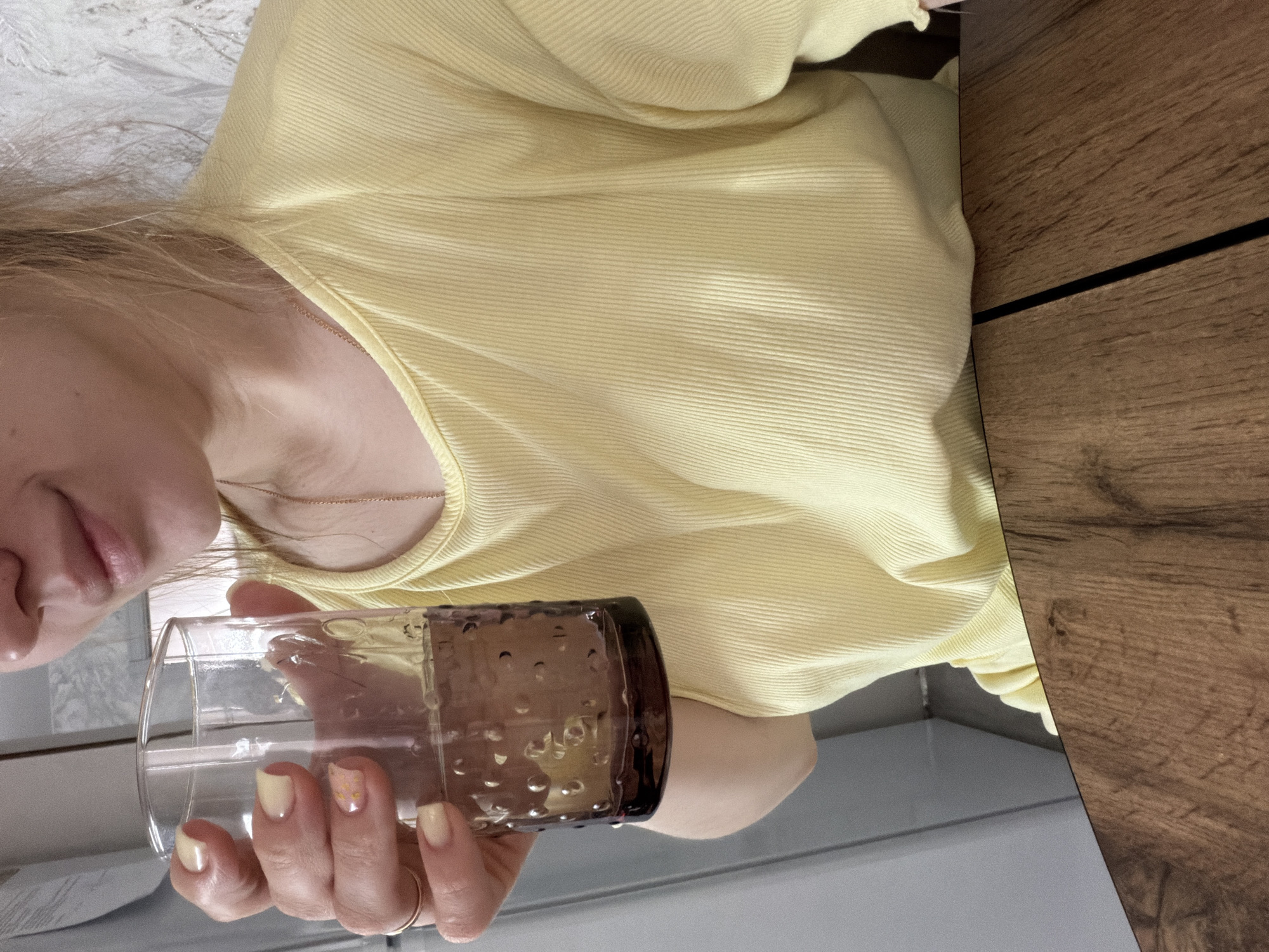 A woman in a ribbed yellow top holds a glass with a dark rim. The lower part of her face is visible, showing her lips. She wears a gold ring on her finger. Her nails are painted in a pale yellow color, with one nail featuring glittery details. The background shows a section of a wooden surface and a light-colored wall or countertop. The location, Reshetnikovo, Russia, cannot be confirmed from the image.