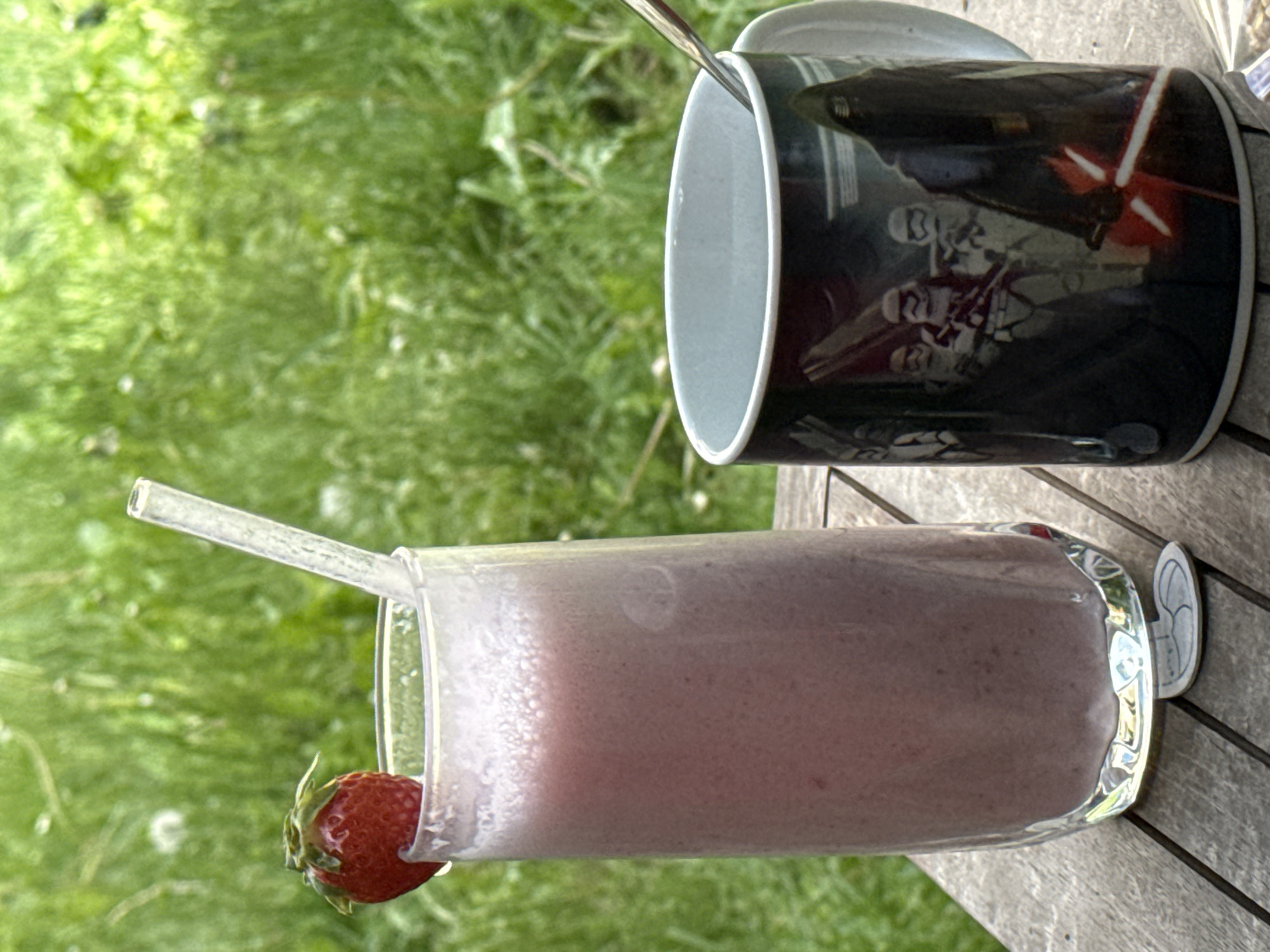 A strawberry smoothie with a clear straw sits in a clear glass alongside a black and white coffee mug adorned with Star Wars imagery on a wooden table, likely outdoors given the green grassy background. The precise location within Sofia, Bulgaria, cannot be determined from the visual information.