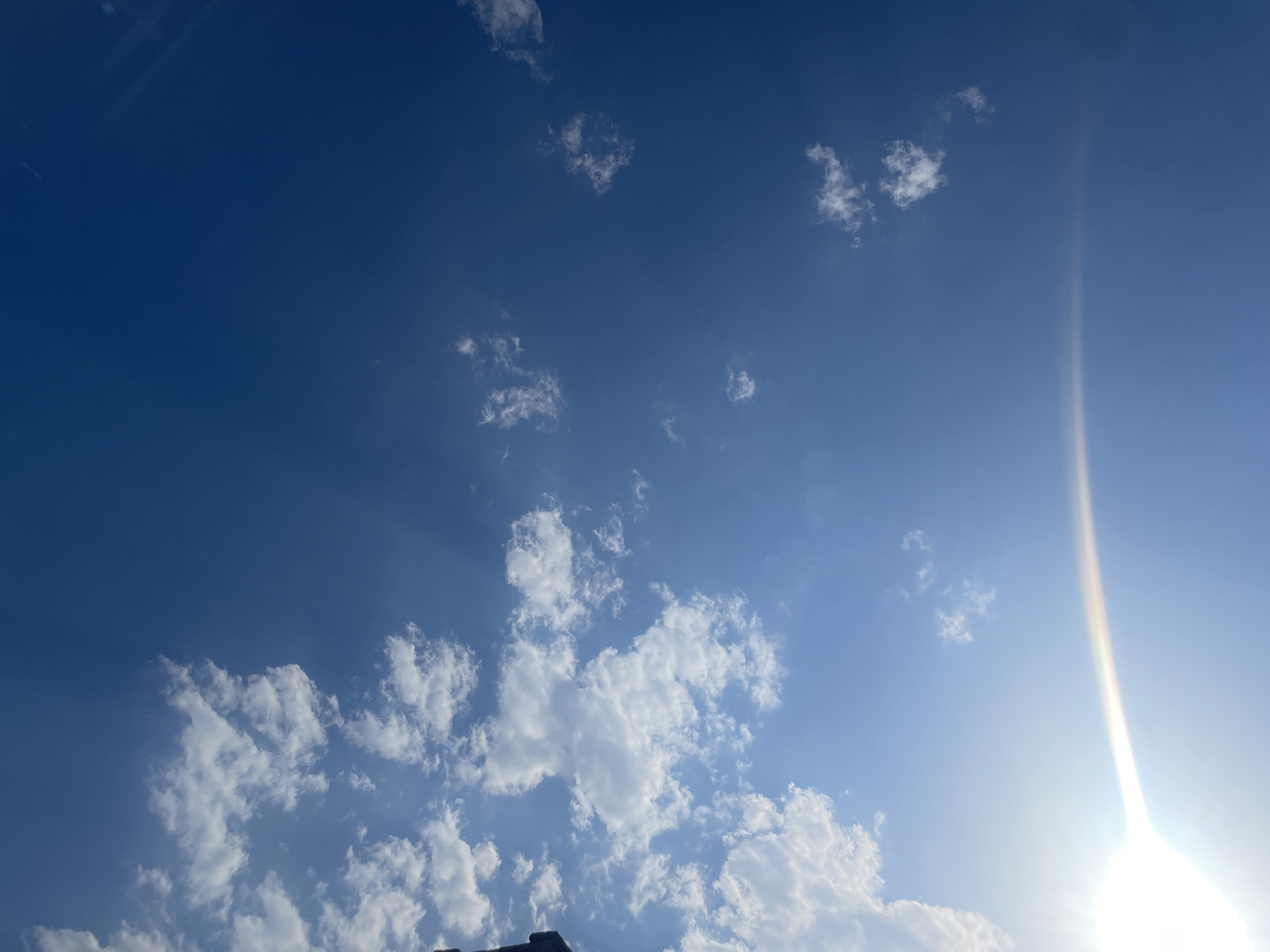 A bright blue sky with scattered cumulus clouds is visible. The sun is setting on the right side of the image, creating a bright glare and a rainbow-like effect. The bottom of the image shows the dark silhouette of a building, possibly a roofline. The scene is devoid of people and any discernible activity. The location is not specified beyond "Unknown, Unknown."