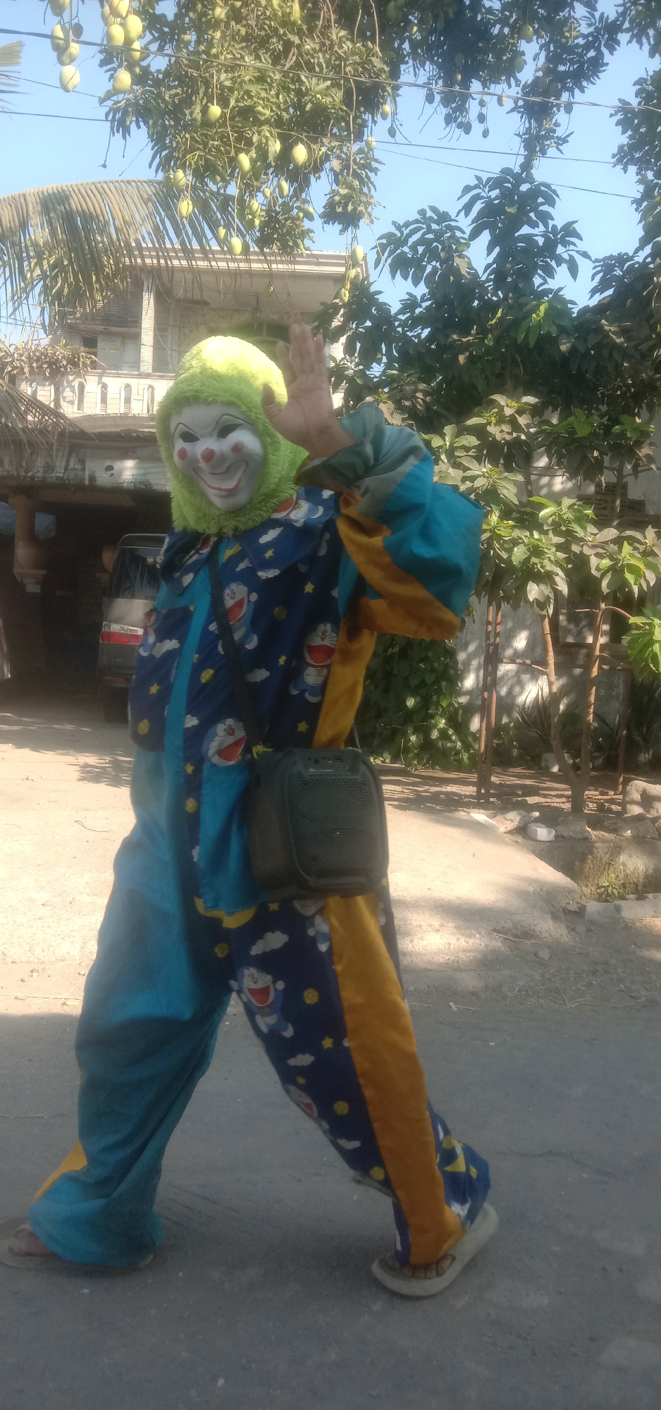A person in a clown costume with a green furry headpiece and white mask with red accents is standing on a paved surface outdoors. The costume is predominantly blue with yellow accents and features a repeating pattern of the character Doraemon. The clown is wearing a black pouch slung across their chest. The person's right hand is raised in a waving gesture. Behind the clown, a light-colored building with a distinctive architectural style is visible, along with trees bearing fruit, likely mangoes, and other foliage. The sky is clear and blue. The scene appears to be located in the city of Banjar Dowo, Indonesia.