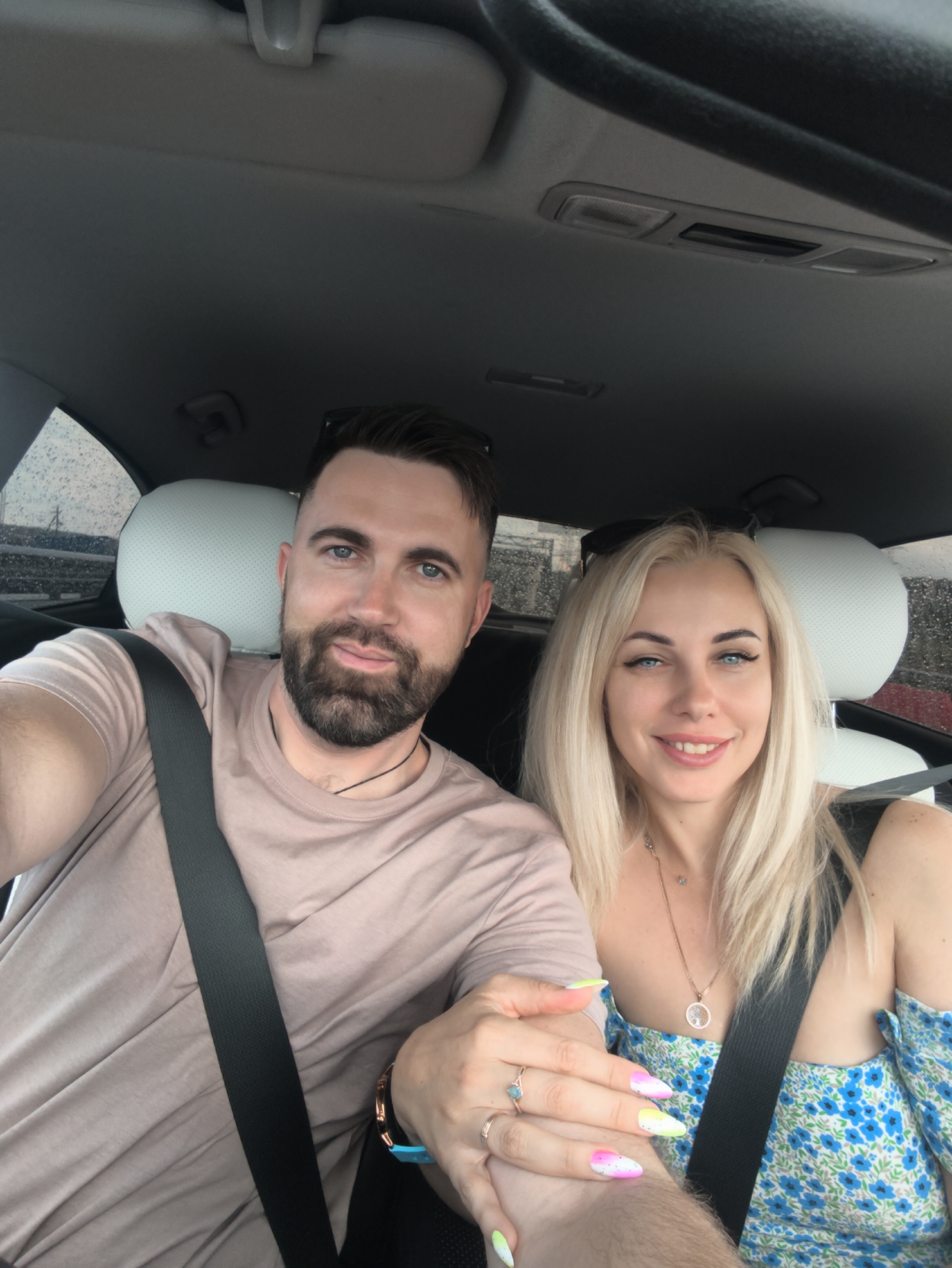A man and a woman are in the front seats of a car. The man is on the left, wearing a light brown t-shirt and a black seatbelt. He has a beard and short dark hair. The woman is on the right, wearing a blue floral dress and a black seatbelt. She has long blonde hair and is wearing a necklace. They are both looking at the camera and smiling. The background shows the interior of the car, including the headrests and the ceiling. The woman's left hand is on the man's arm, and she has colorful painted nails. The man's right hand is holding the woman's left hand. The scene appears to be during the day, with some rain visible on the car windows. The location is not identifiable as Kerch, Ukraine or any other specific city.