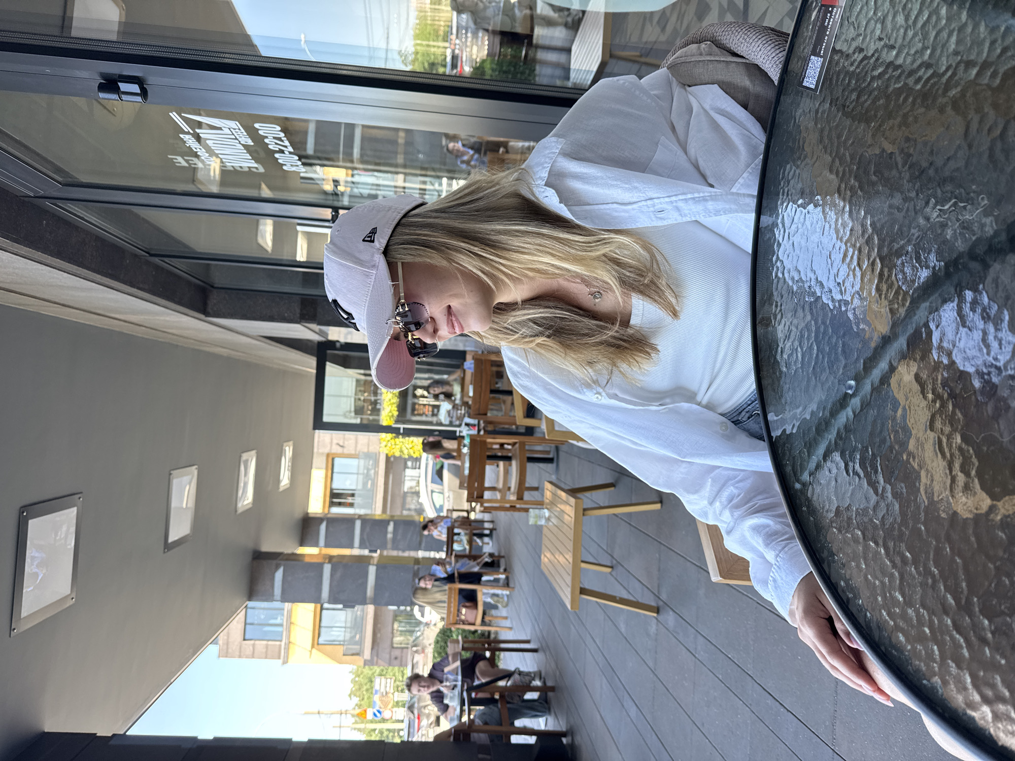 A young woman with blonde hair, wearing a white cap, sunglasses, and a white open button-up shirt over a white tank top, sits at a glass-top table on an outdoor patio. She is looking to her right. The patio has a grey tiled floor and is lined with tables and chairs. In the background, people are seated at other tables, and a building with large windows and columns is visible. The scene appears to be in a city during the daytime, with a bright sky.