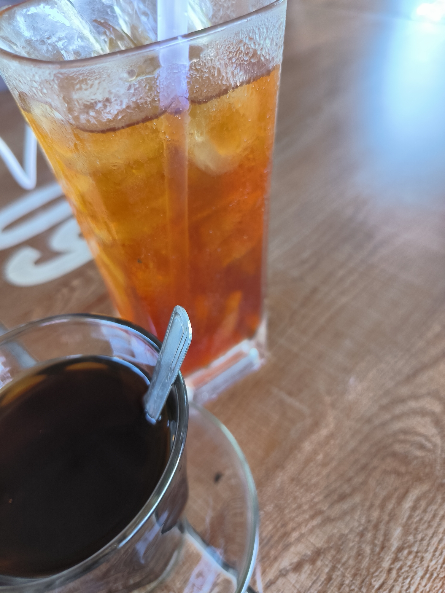 A close-up shot on a wooden surface shows two beverages: a glass of ice tea and a cup of black coffee, with a spoon in it. The ice tea, in a tall clear glass, has condensation on the outside. The coffee is in a clear glass cup on a clear saucer. The setting is likely a cafe in Matang, Indonesia. There is a light source creating highlights on the table.