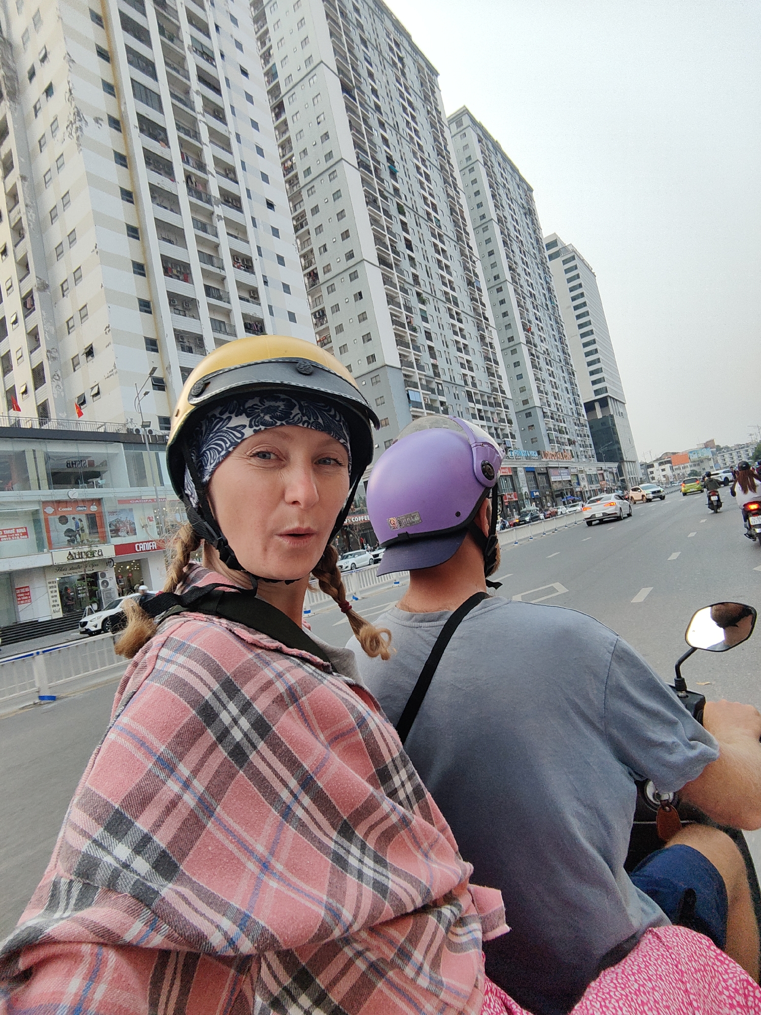 A low-angle, medium shot shows a light-skinned woman with blonde pigtails in a yellow helmet and plaid shirt, as a passenger on a motor scooter in Hong Gai Ward, Vietnam. She faces the camera with a surprised expression. The scooter is driven by a man with a light-skinned neck in a purple helmet and gray t-shirt. The scooter is in traffic on a multi-lane road. Modern high-rise apartments of neutral colors line the road. Several cars and scooters are also on the road. Business storefronts are visible on the left. The light is bright but the sky is overcast.