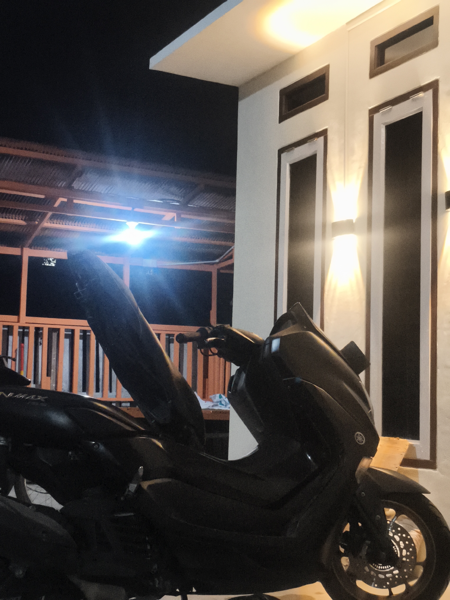 The image shows a dark-colored Yamaha NMAX scooter parked outdoors at night, illuminated by artificial lights. The scooter is positioned in front of a modern building with white walls and two tall, narrow windows. Above the windows, there are smaller, horizontally oriented windows. The building is lit by two wall-mounted light fixtures that cast a warm, upward glow. To the left of the building, a wooden structure with a corrugated roof is visible, also illuminated by a bright light that appears to be a headlight from the scooter itself, causing a strong blue glare. The ground is not clearly visible due to the low light conditions and the scooter's placement. The overall impression is that of a residential or commercial property in an urban or suburban setting. While the location is stated to be Samarinda, Indonesia, there are no specific visual cues within the image that definitively confirm this location.