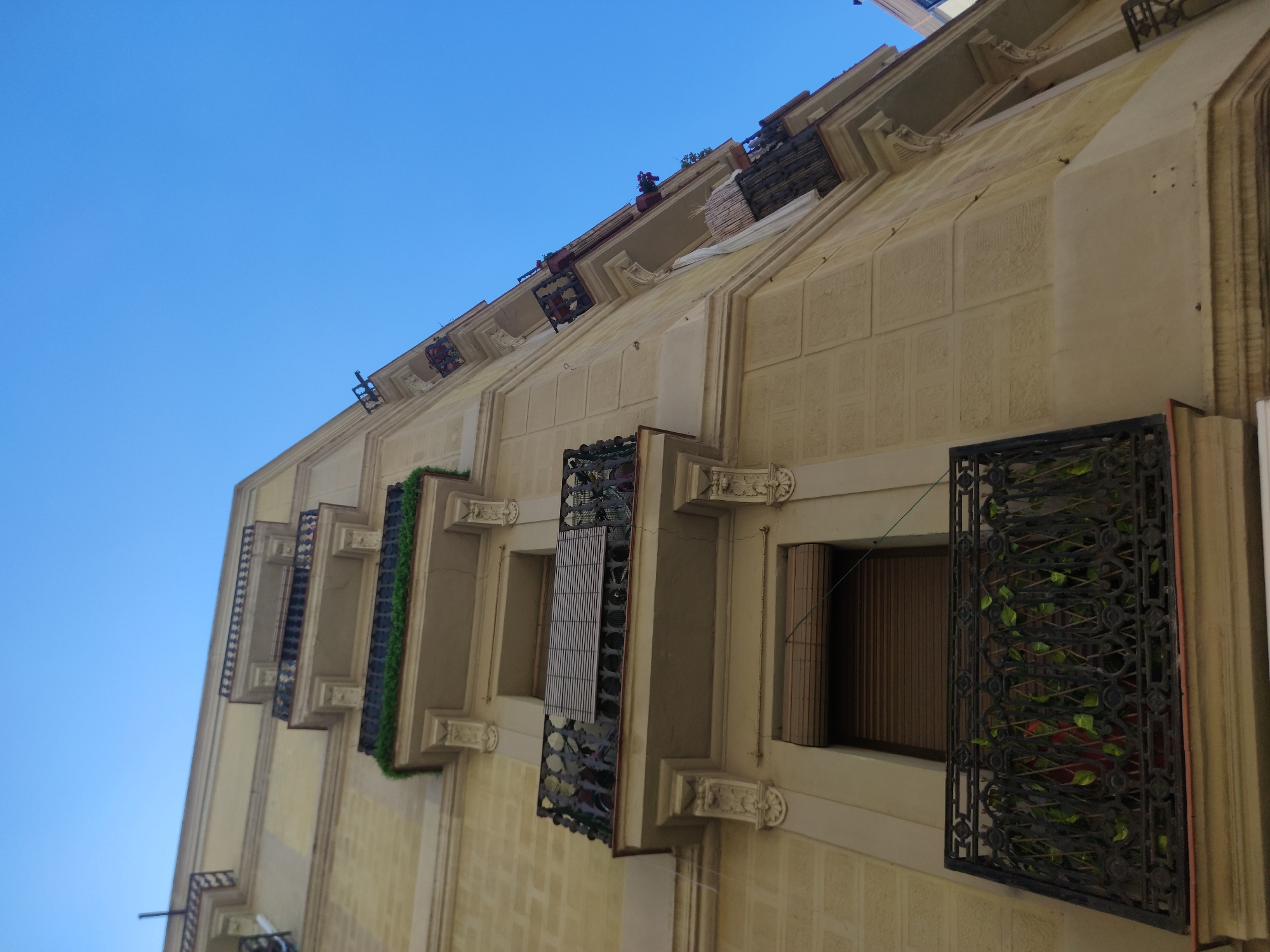 Captured from a low-angle perspective, the image showcases the architectural detail of a building's exterior. The structure is characterized by its tan façade, punctuated by a series of balconies with decorative ironwork. Some balconies are adorned with vegetation. Windows are set into the facade, with one partially obscured by closed blinds. Above the balconies, the building's design incorporates geometric ornamentation. A clear, blue sky serves as the backdrop, emphasizing the building's vertical rise. The location is in the city of Unknown, Unknown.