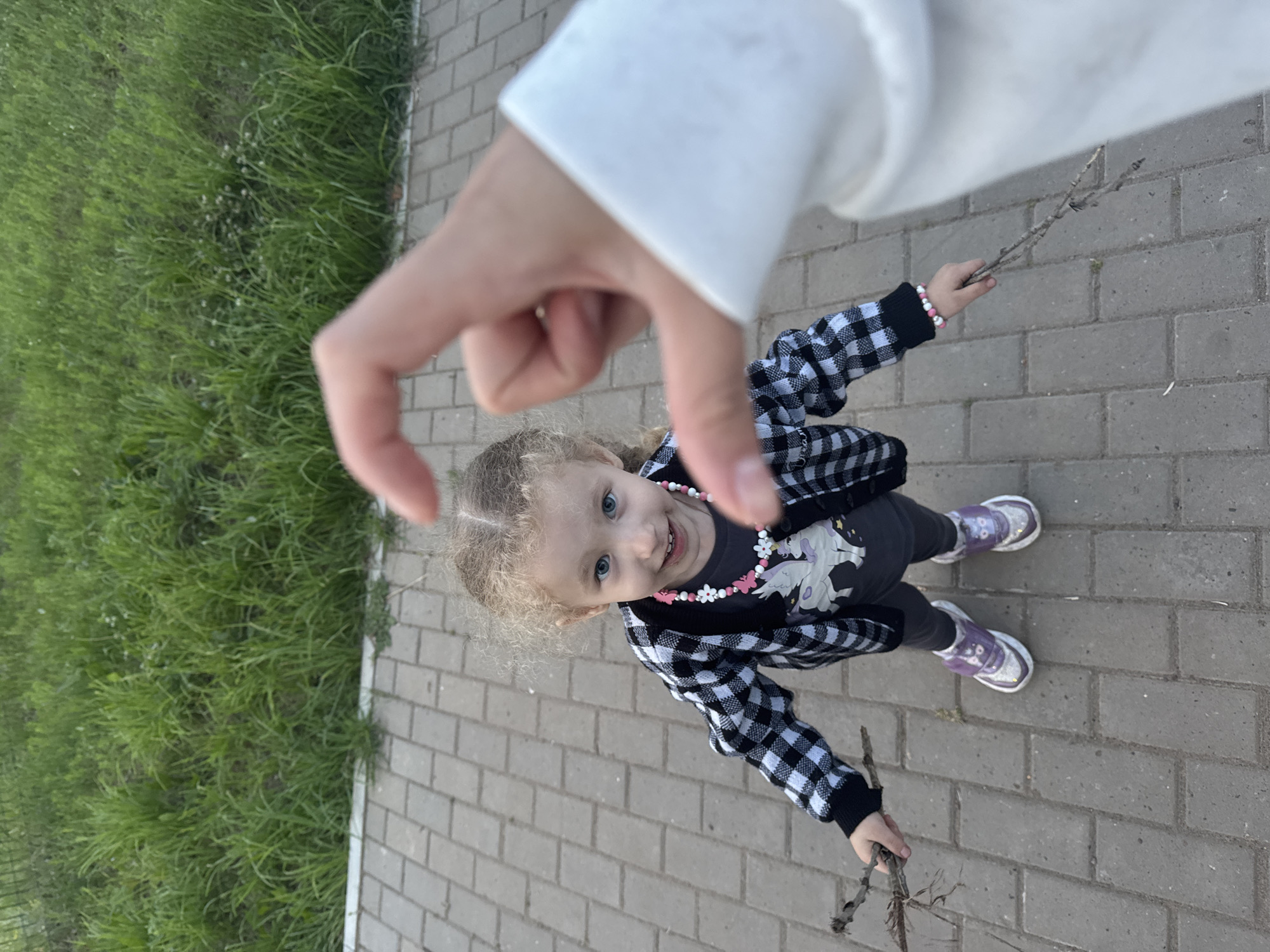 The media file shows a young girl with blonde, curly hair and blue eyes, looking up at the camera with a smile. She is wearing a black and white checkered jacket, dark pants, and purple sneakers. Around her neck, she has a necklace with pink and white flower-shaped beads. She is holding a small stick in her right hand. In the foreground, a hand is making a heart shape, seemingly framing the girl. The girl is standing on a paved walkway made of grey bricks. To the left of the walkway is a patch of green grass with some small white flowers. The image appears to be taken outdoors during the daytime, likely in a park or a similar public area. The overall mood is playful and affectionate. There is no visible text in the image. The location context provided is Chaikovsky, Russia.